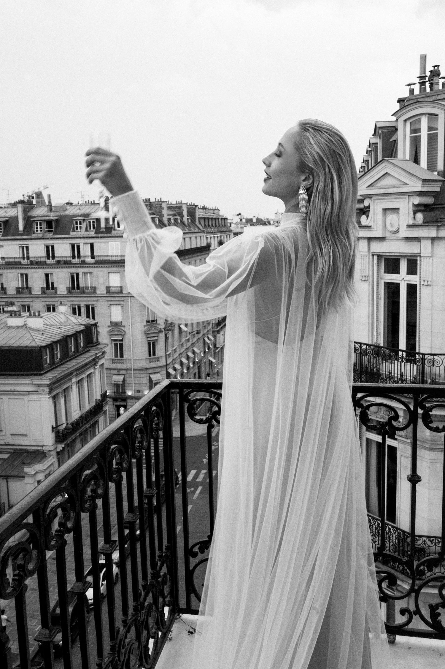 Blonde bride in flowing white gown raises champagne glass on ornate Parisian balcony in black and white.