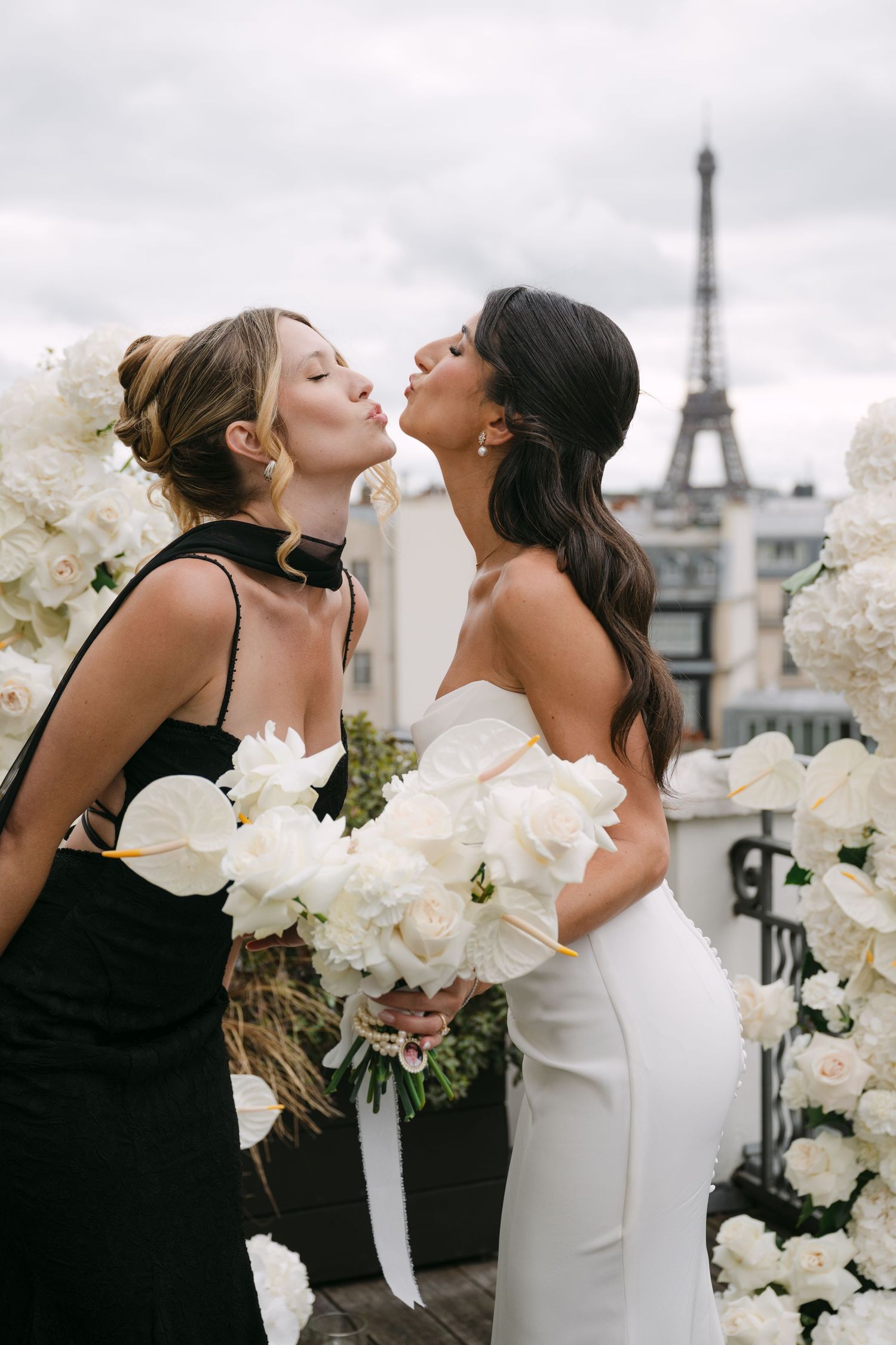 Bride and bridesmaid share a playful kiss pose before white floral arch with Eiffel Tower visible.