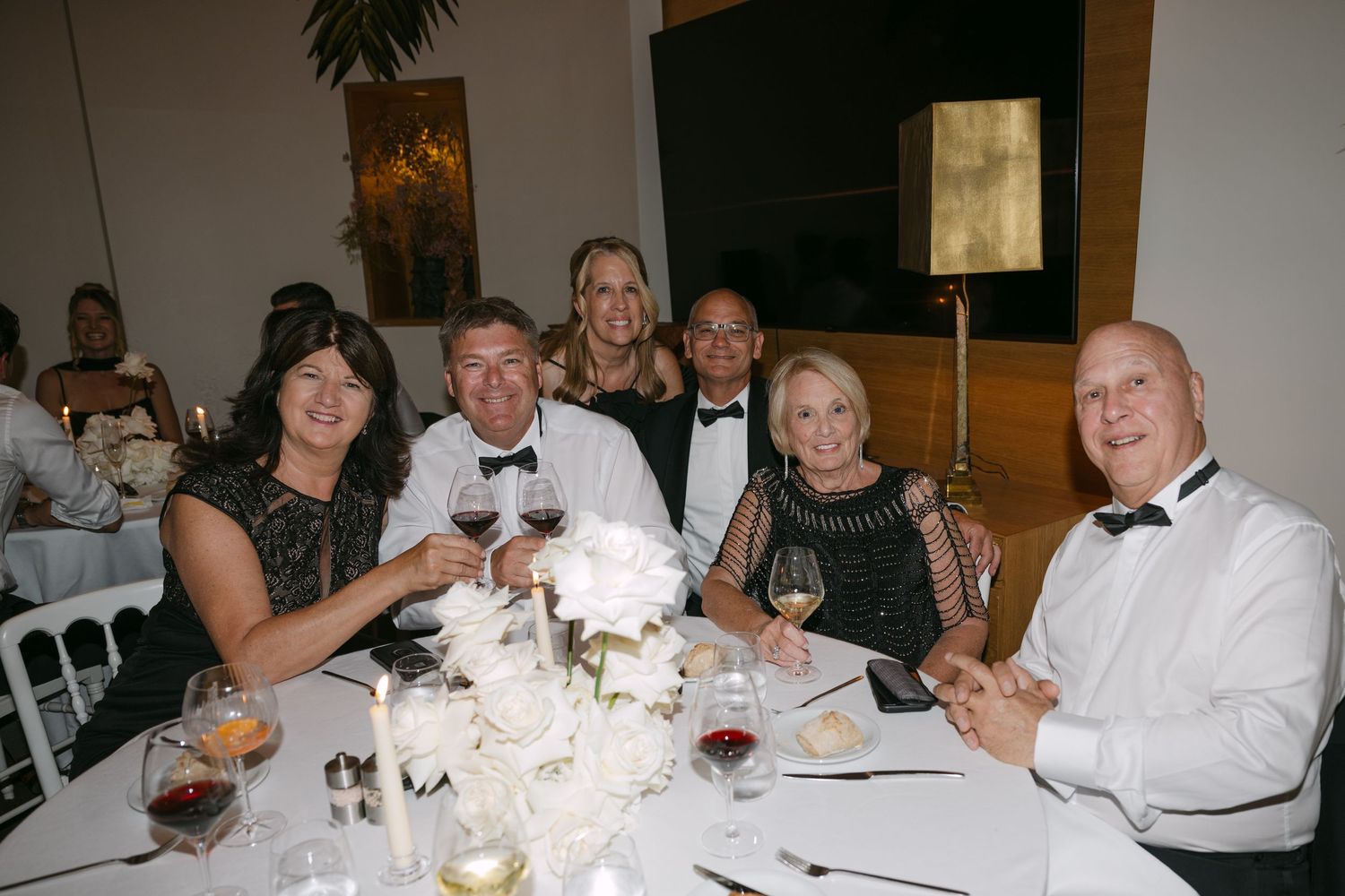 Group of six guests in formal attire toasting at a wedding reception dinner table.