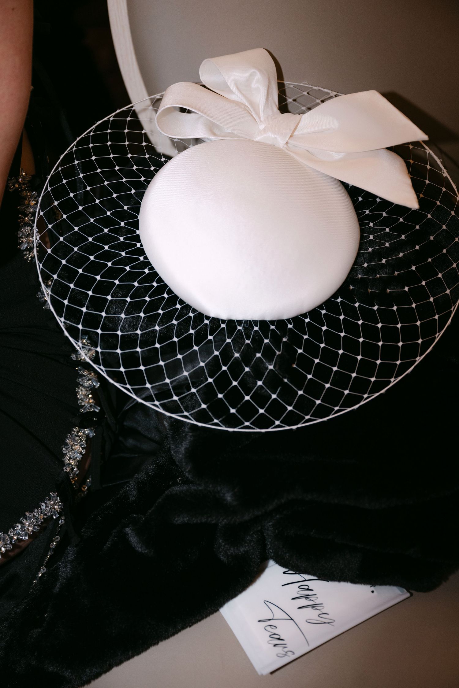 White pillbox bridal hat with bow and birdcage veil resting on black fabric with calligraphy card.