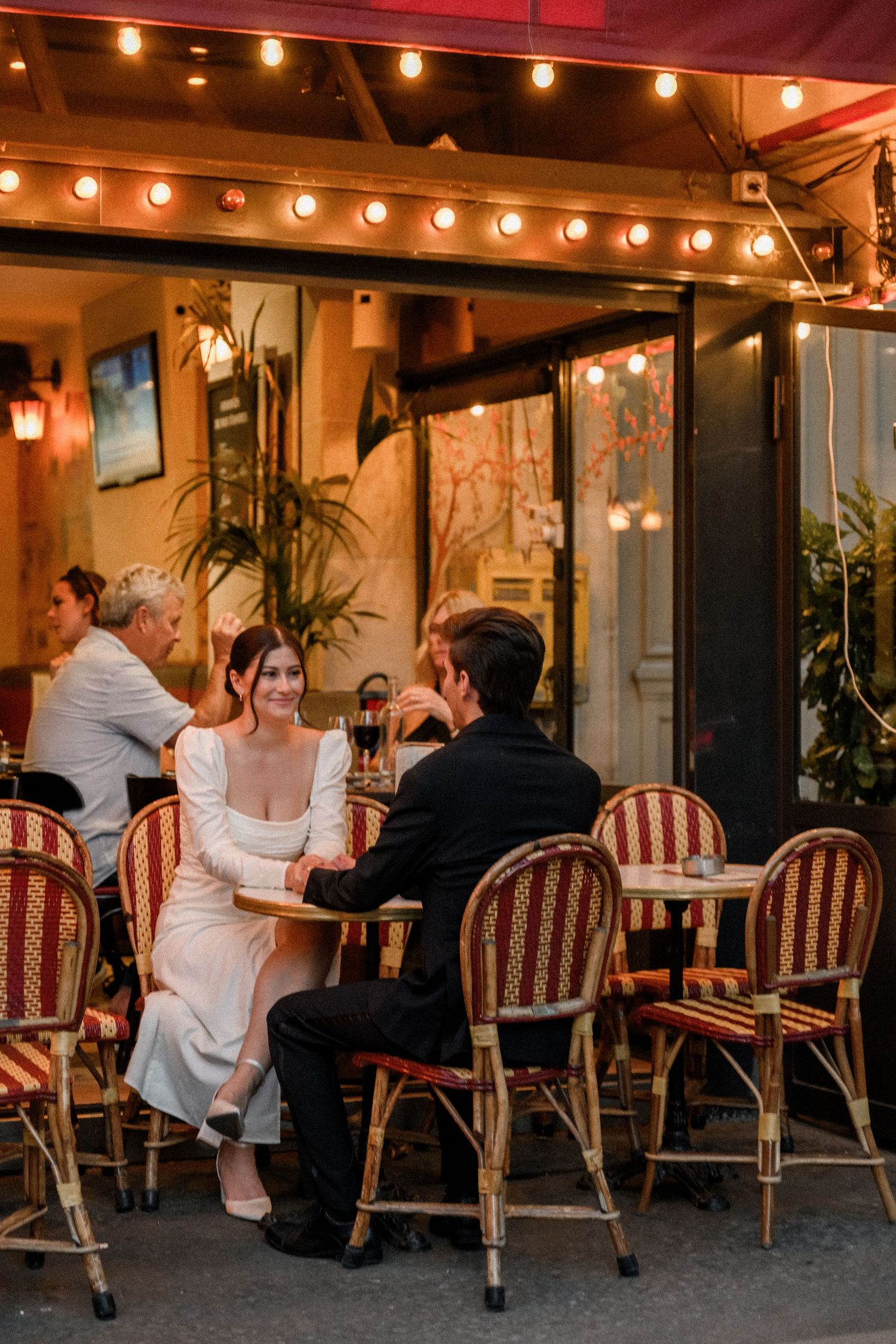 A bride and groom share an intimate moment at a Parisian outdoor cafe with warm string lights.