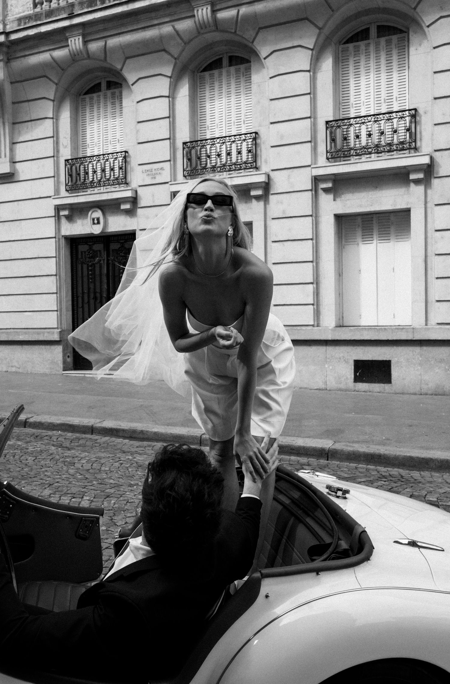 Black and white photo of bride in veil standing in vintage convertible with groom on cobblestone Paris street.