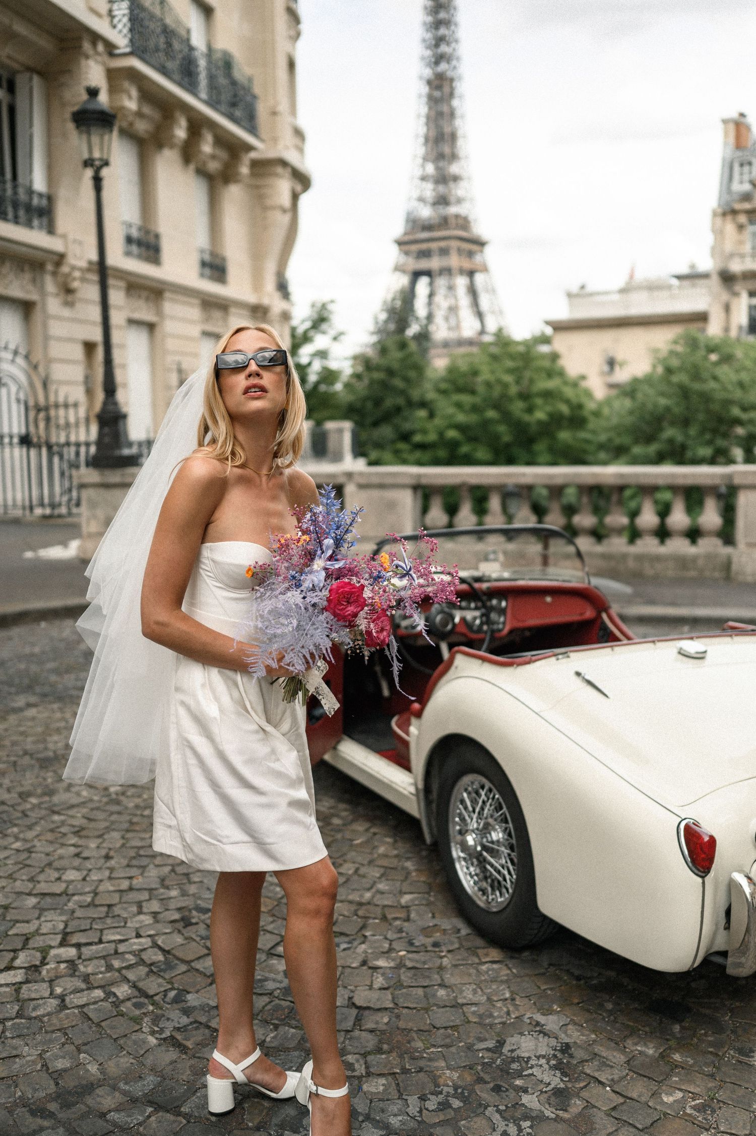 Blonde bride in short white dress and sunglasses holding colorful bouquet beside vintage Triumph car with Eiffel Tower.