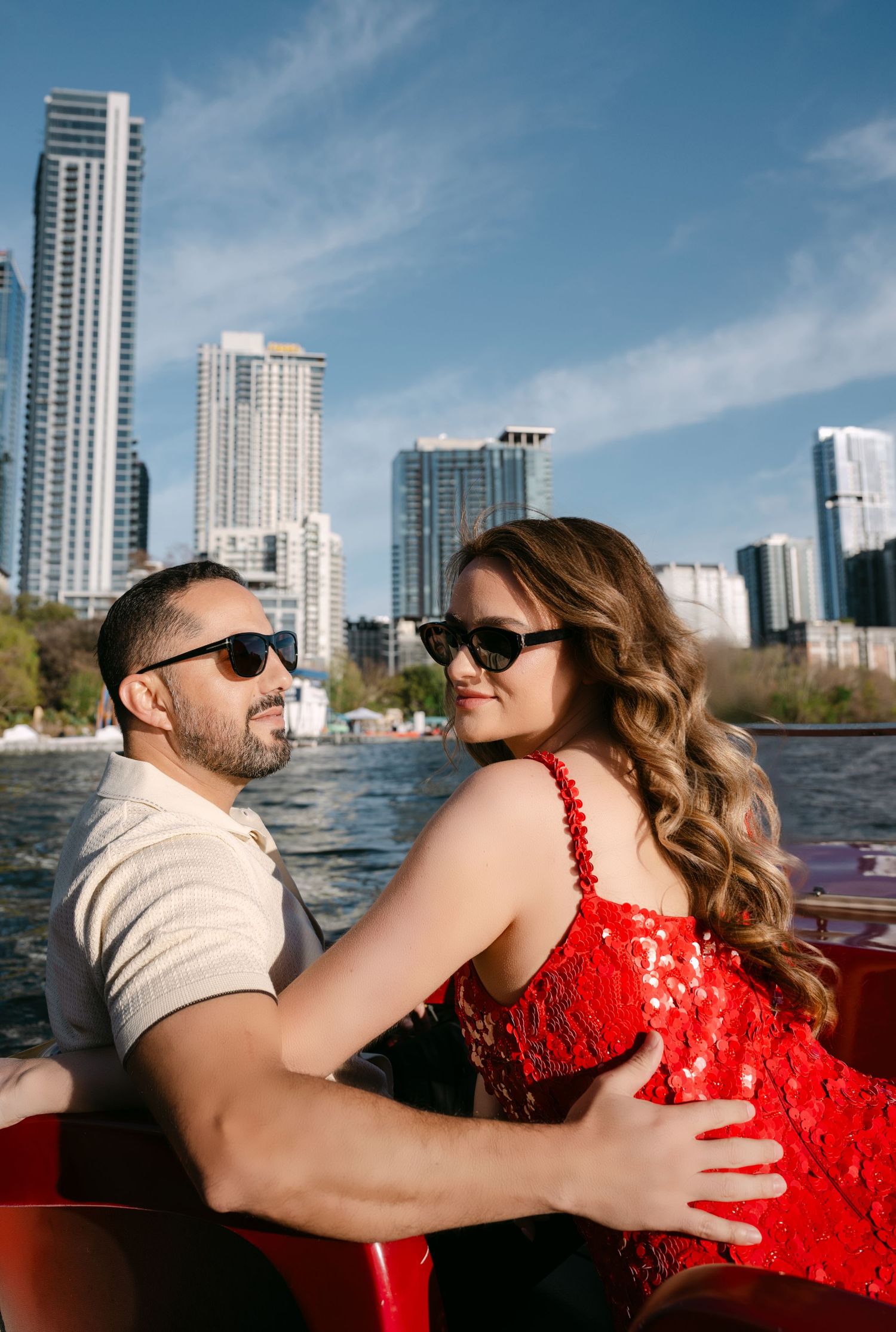 Couple in sunglasses embrace on red vintage boat with city skyline and blue sky in background.
