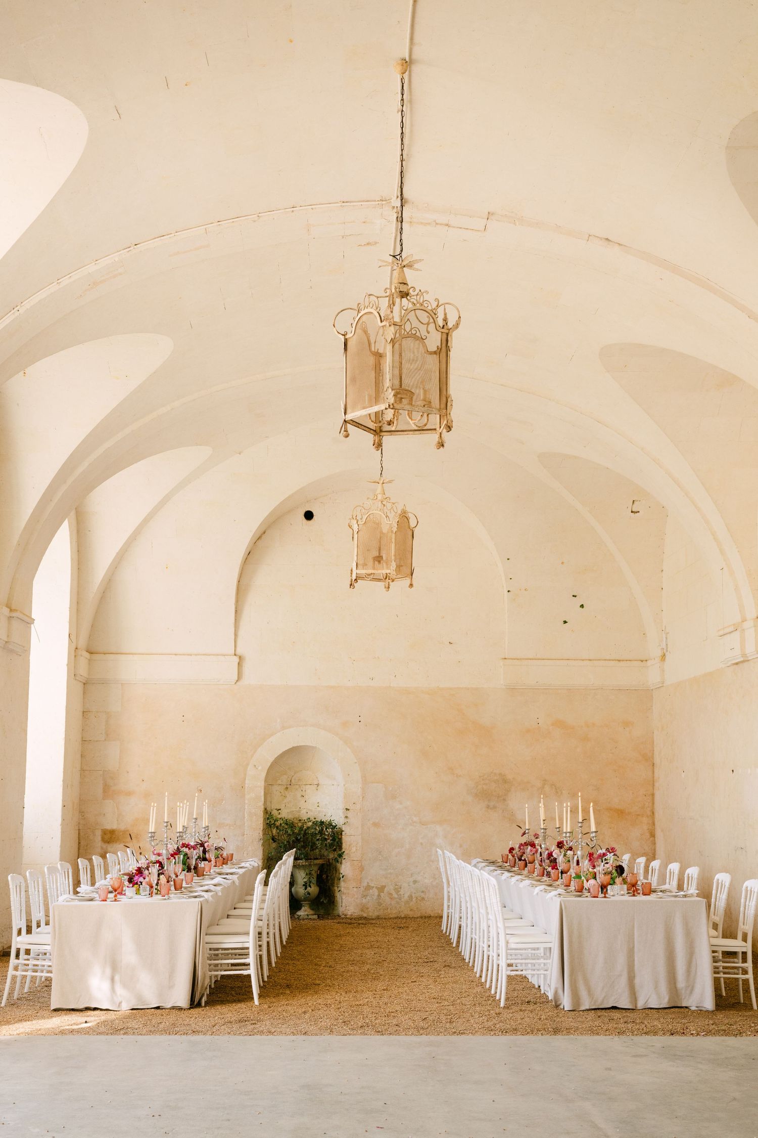 Vaulted stone ceiling wedding reception hall with hanging lanterns and long tables set with pink floral arrangements.