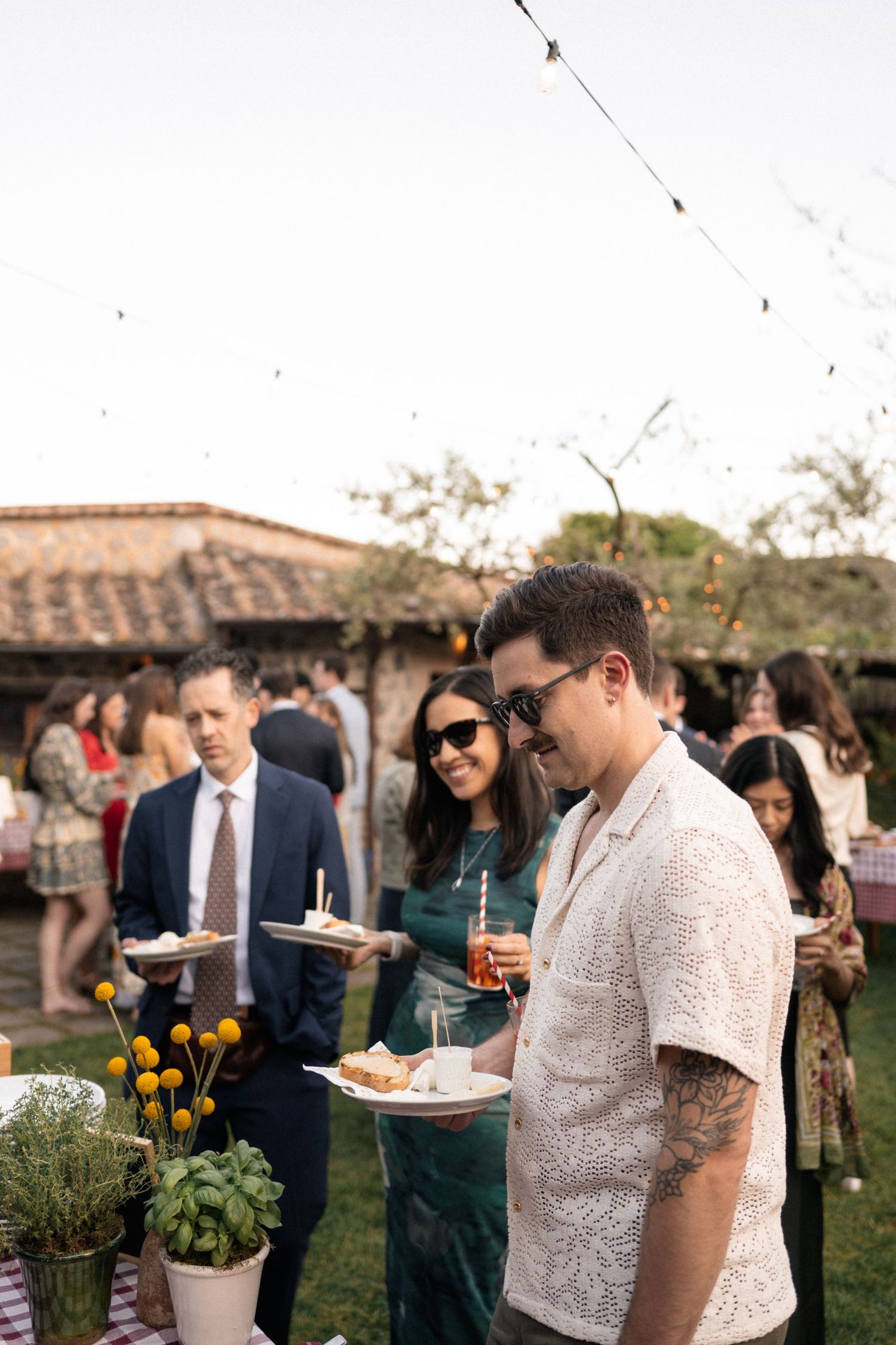 Wedding guests enjoy appetizers and cocktails at a sunny outdoor Italian reception with string lights.
