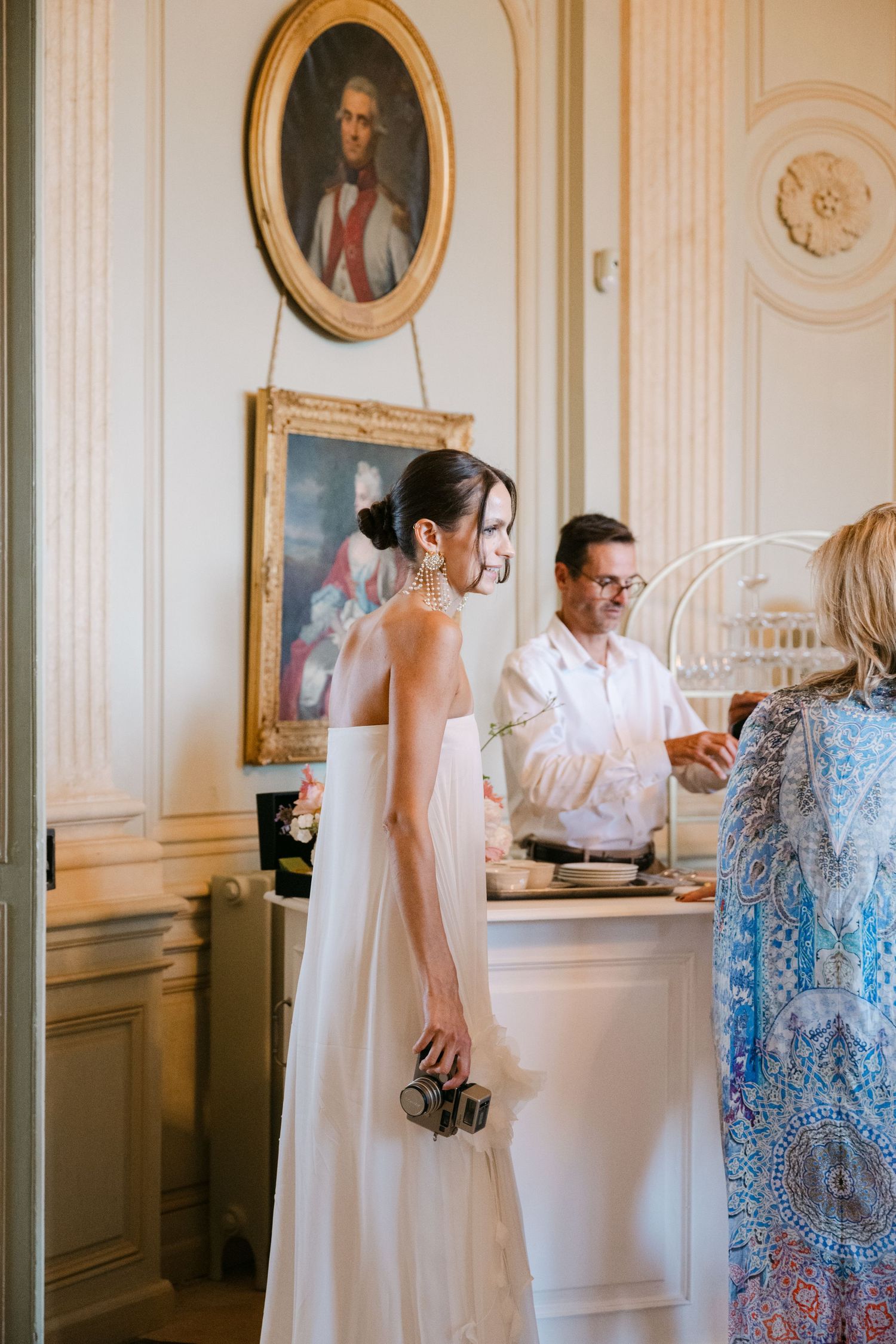 Bride in white gown stands near a buffet table beneath an oval portrait painting in a French chateau.
