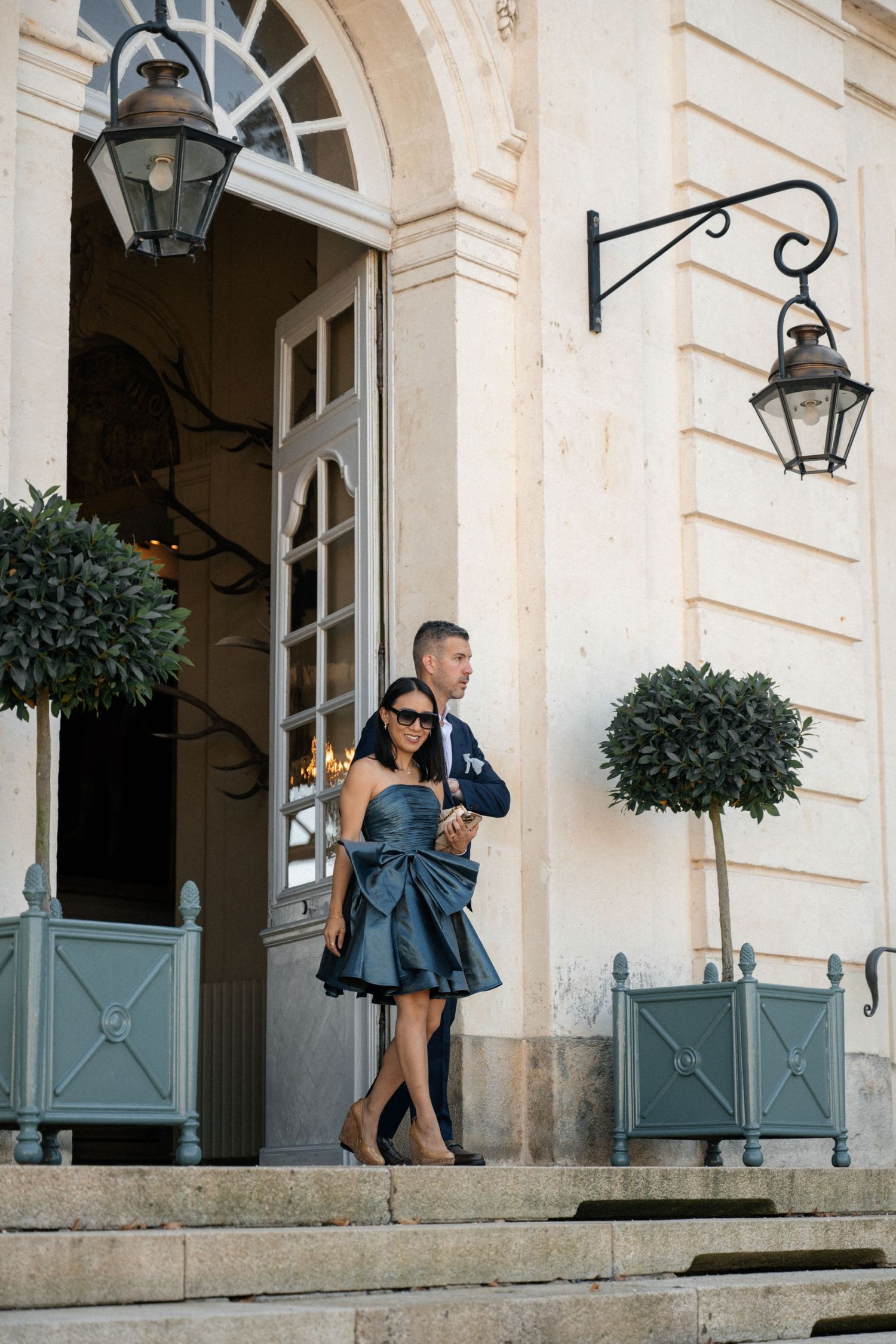 Stylish couple posing on the entrance steps of a classic French chateau with decorative lanterns.