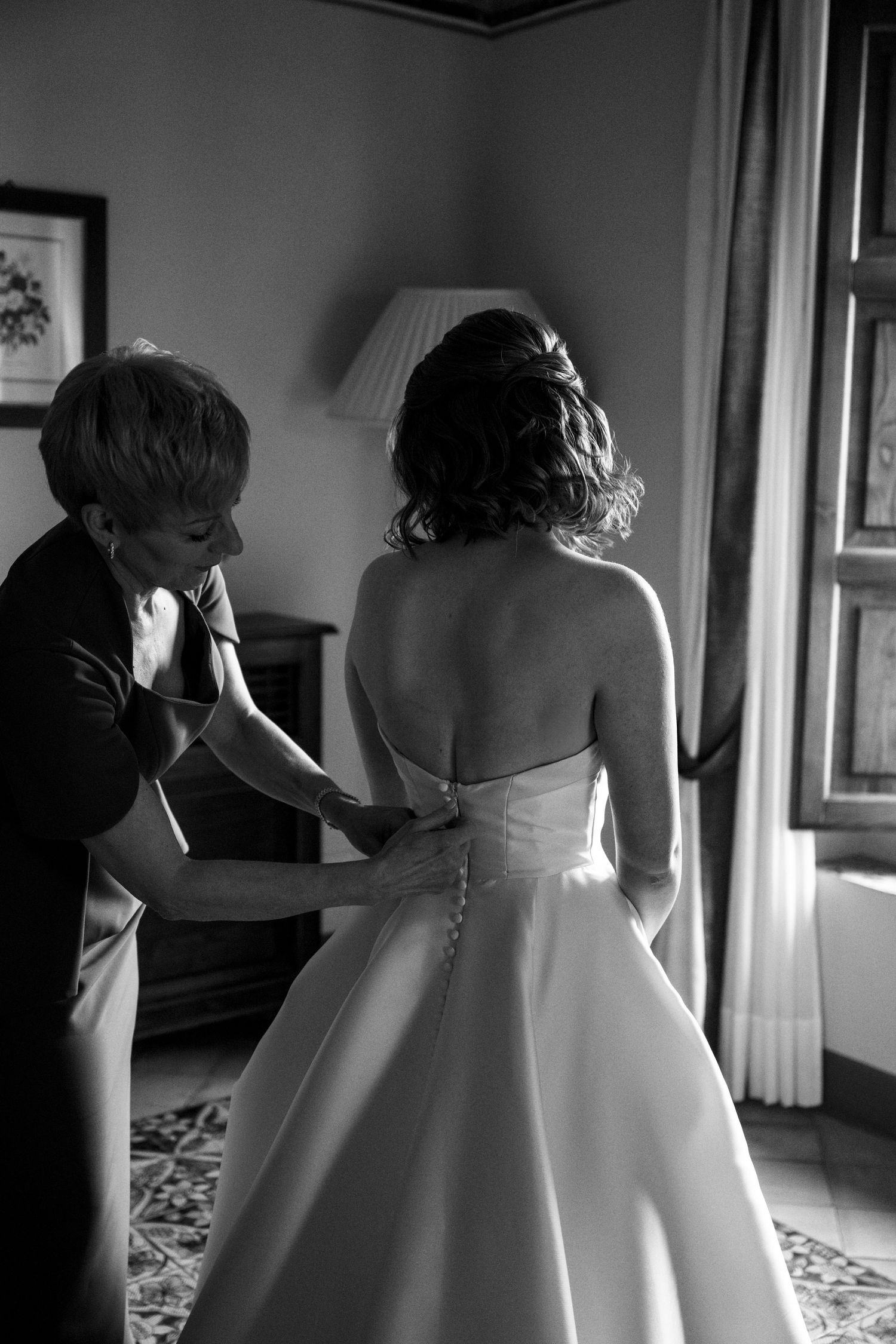 Black and white photo of woman buttoning bride's strapless wedding gown in hotel room.
