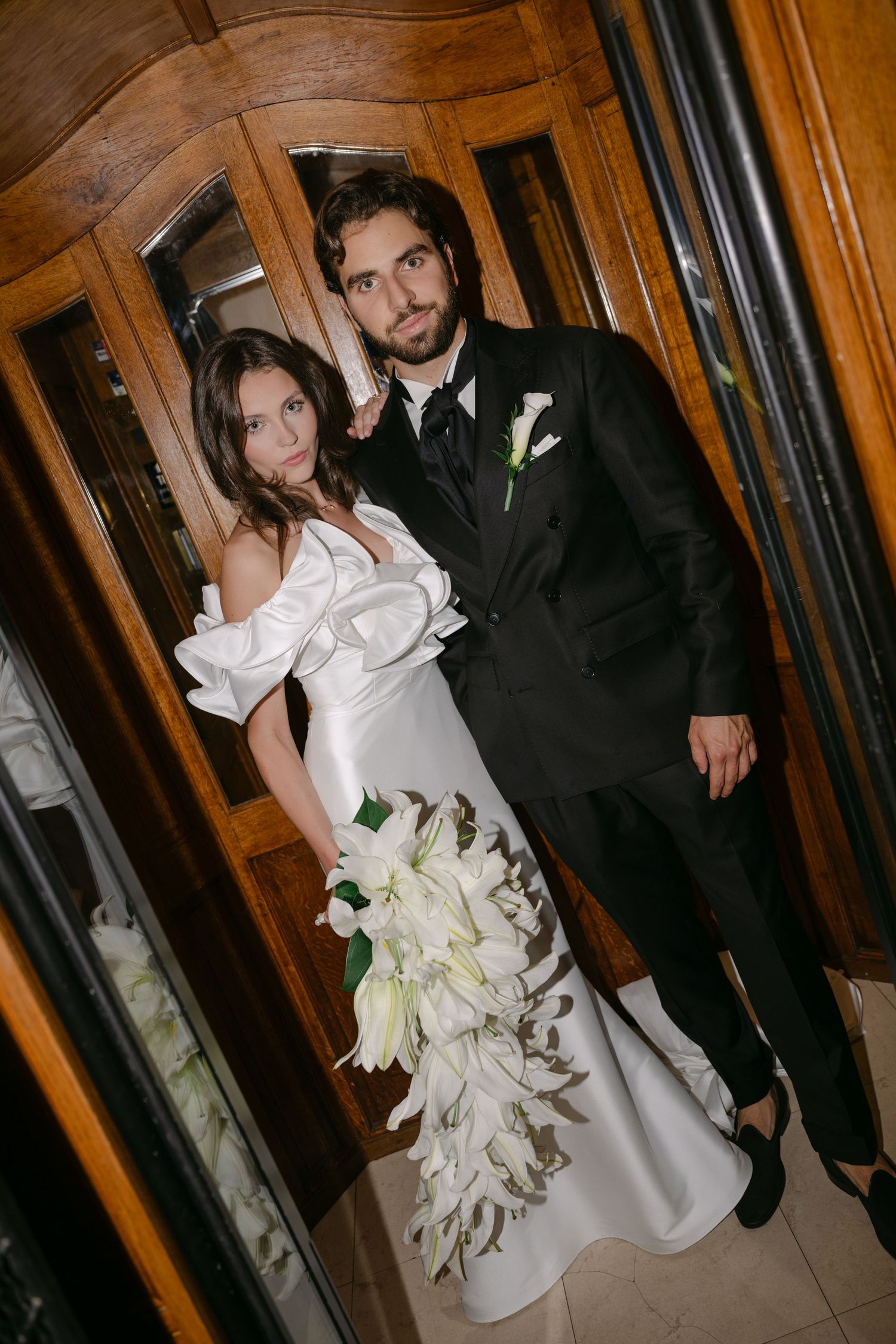 Elegant bride in white off-shoulder gown holding white lily bouquet poses with groom in black tuxedo by wooden doors.