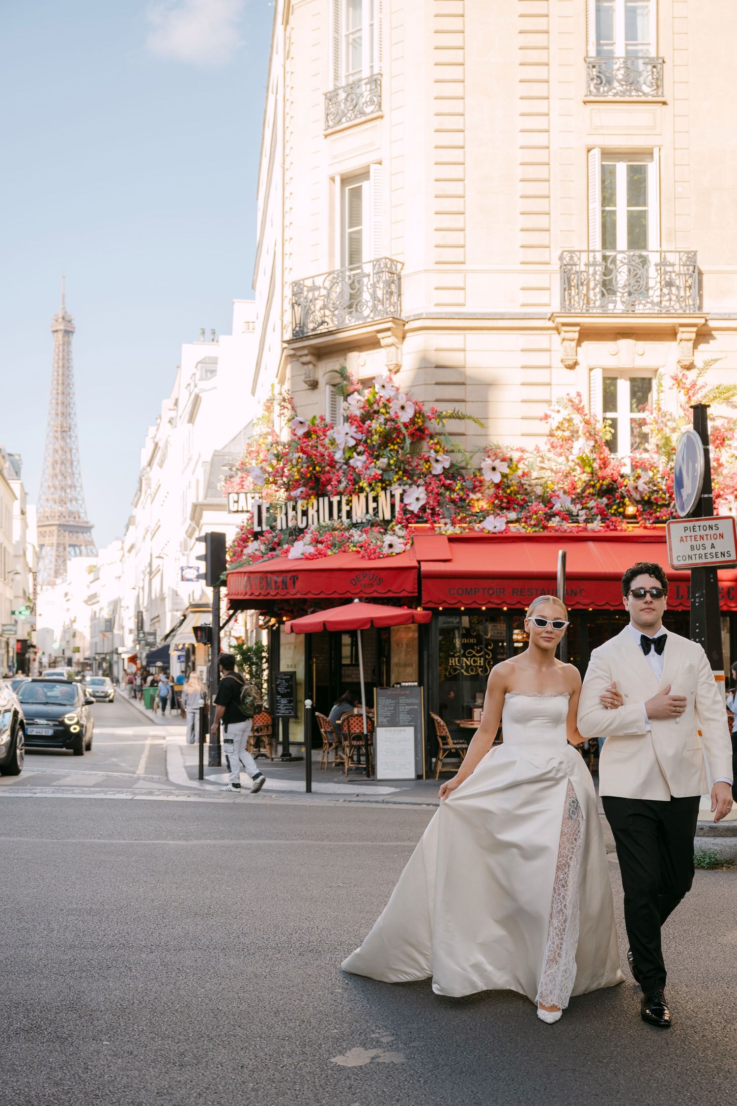Stylish newlyweds walking past a Parisian café with the Eiffel Tower visible in the background.