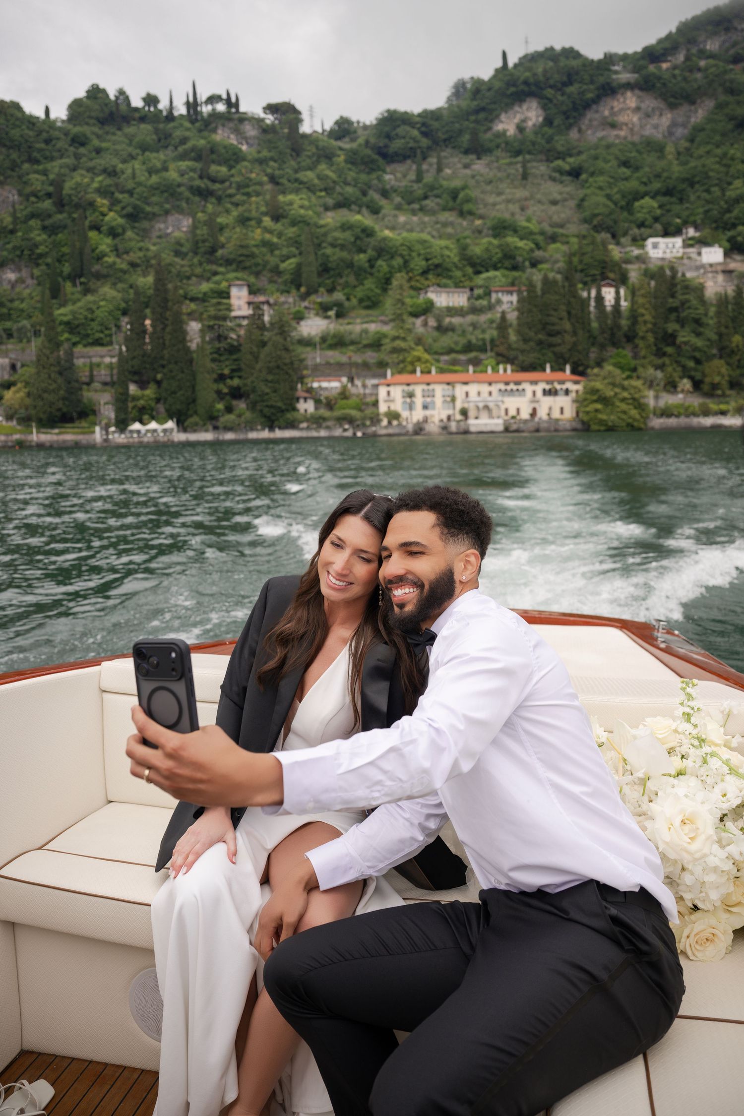 Smiling newlyweds taking selfie on wooden boat with stunning Italian lake and hillside backdrop.