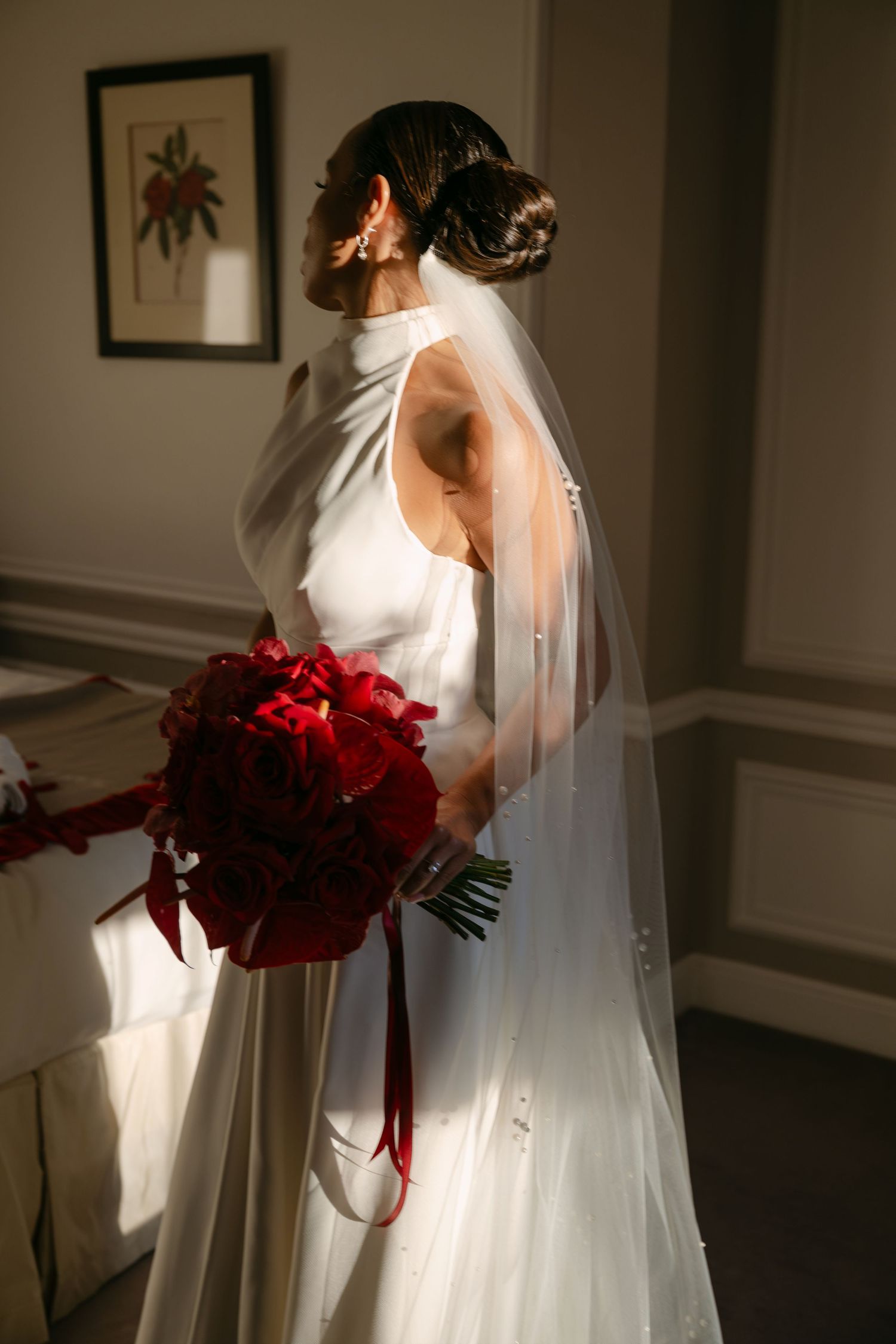 Bride in white halter gown and veil holds red rose bouquet, standing in soft natural light by a window.