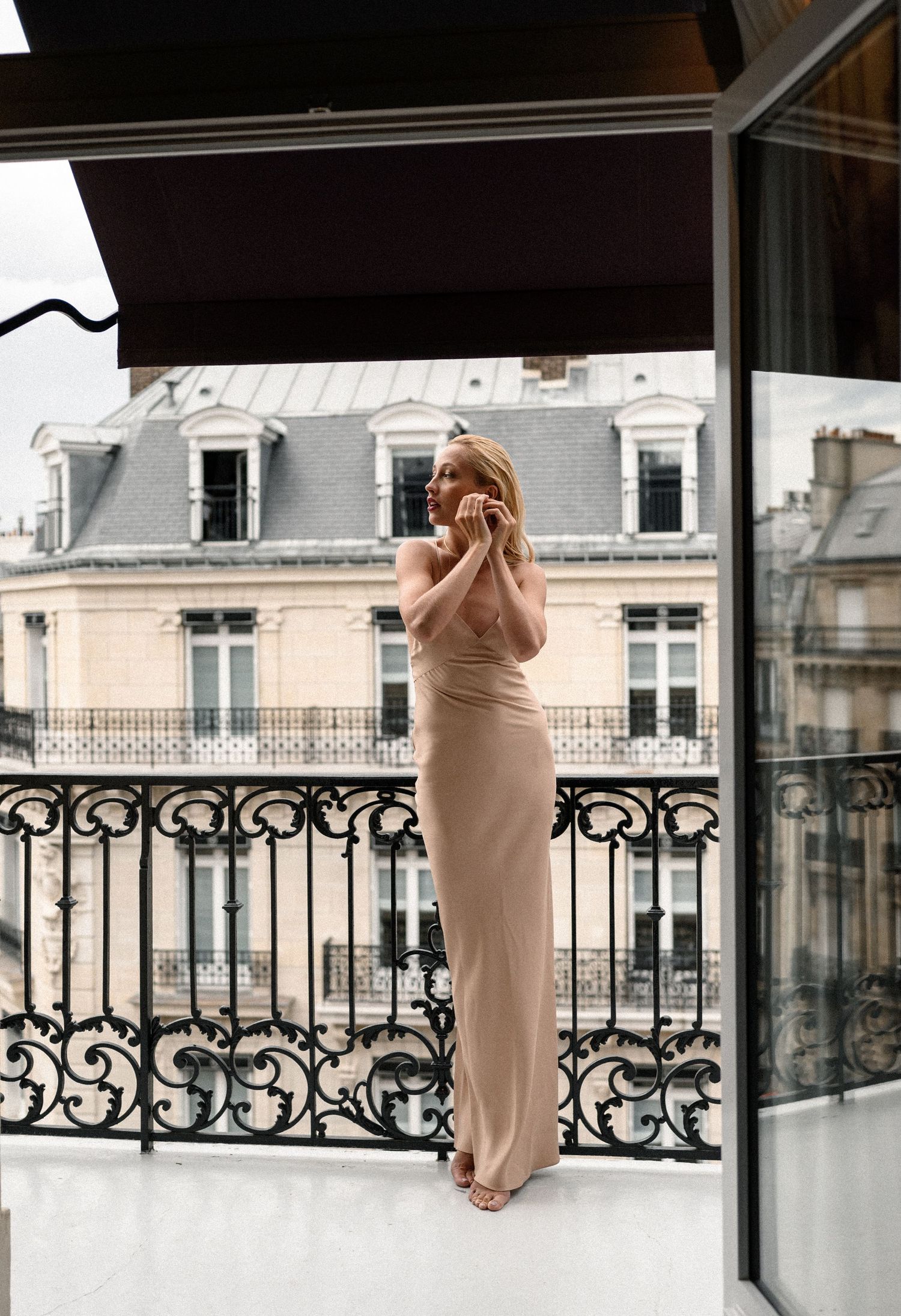 Blonde bride in champagne silk gown standing on luxury Parisian hotel balcony with classic architecture backdrop.