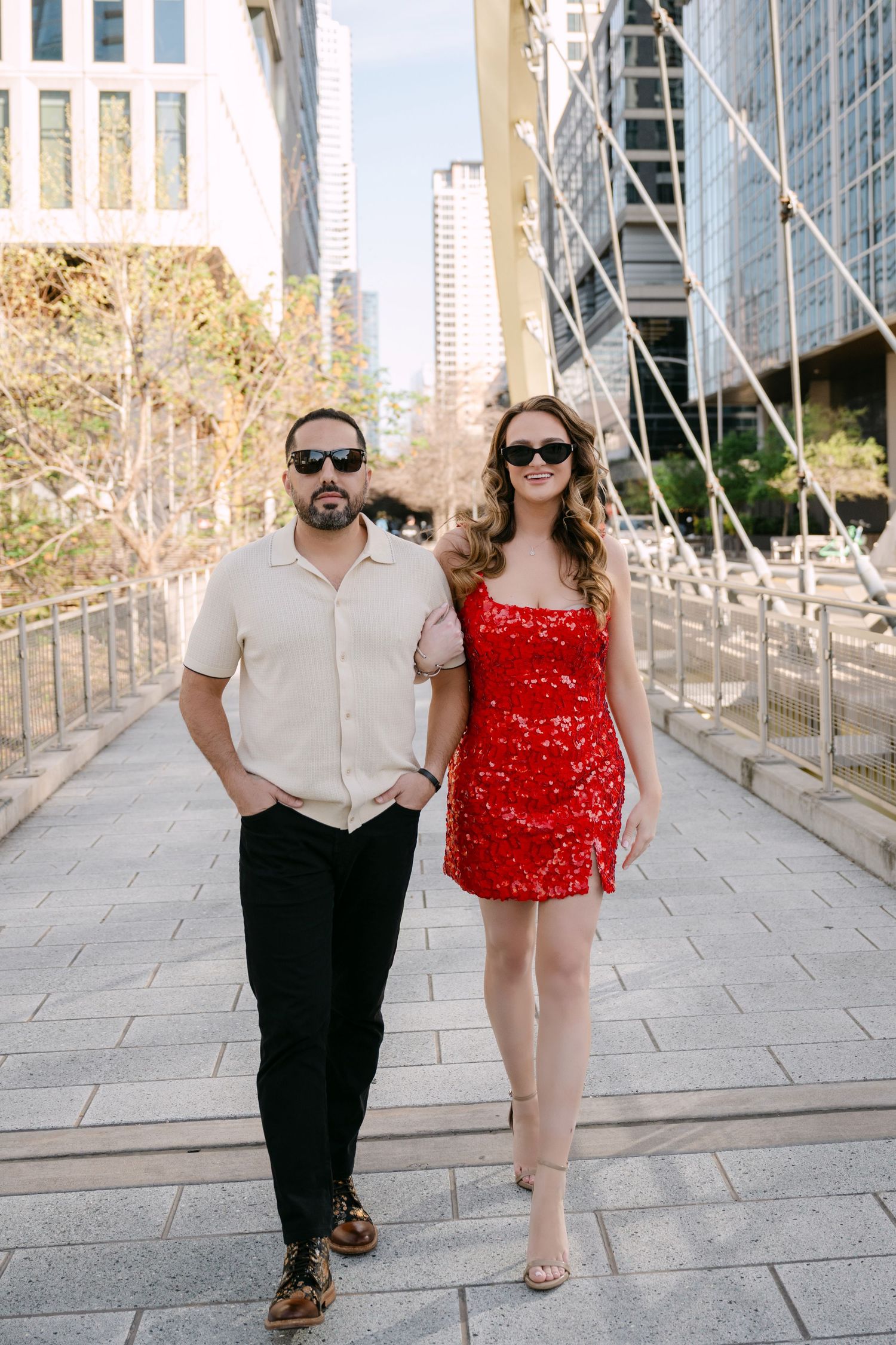 Stylish couple walking down city steps, woman in red sequin mini dress and man in beige shirt with sunglasses.