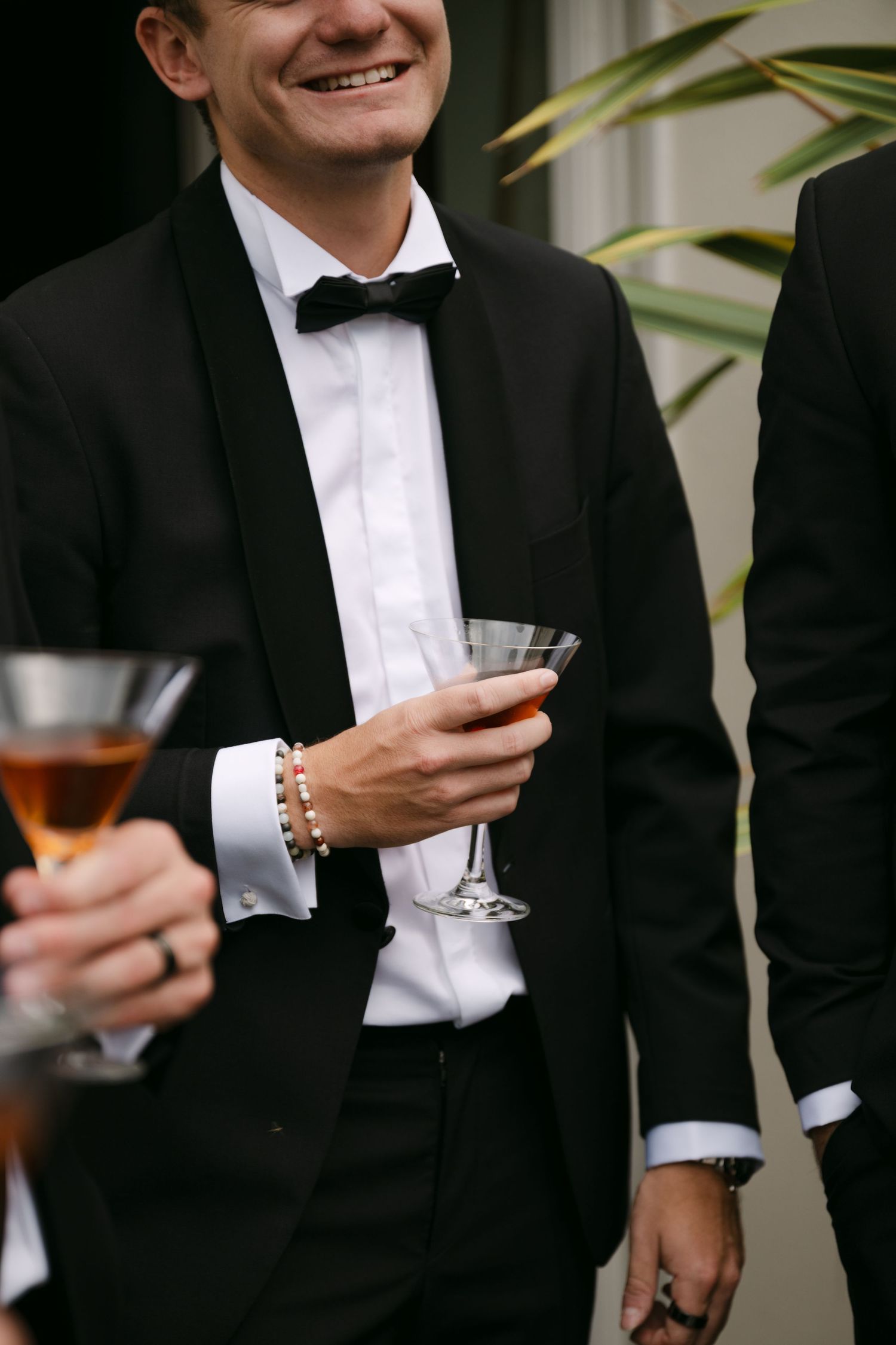 Smiling man in a black tuxedo with bow tie holding a martini glass at an elegant formal event.