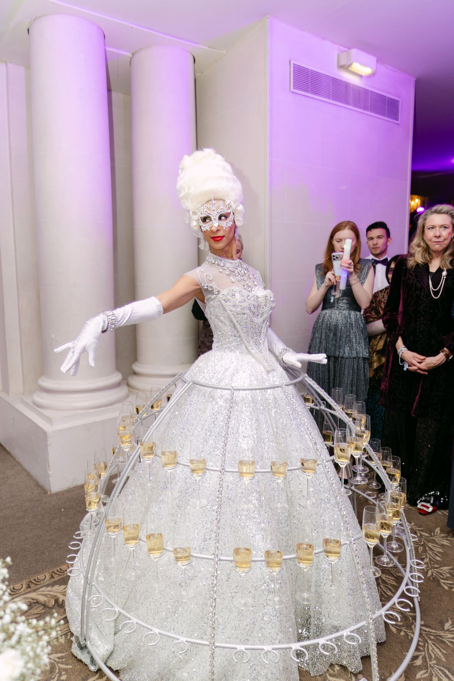 Stunning performer in silver sequined gown with champagne glasses on hoop skirt at an upscale gala.
