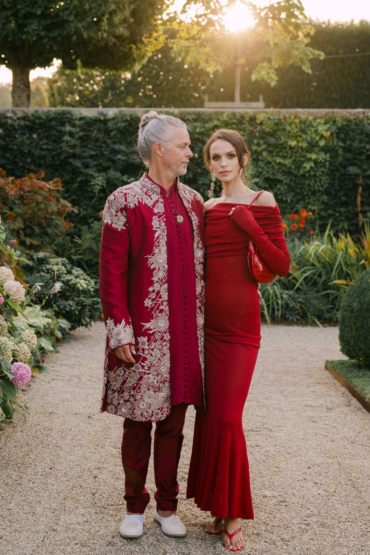 Elegant couple posing in coordinated red formal attire in a sunlit garden setting.