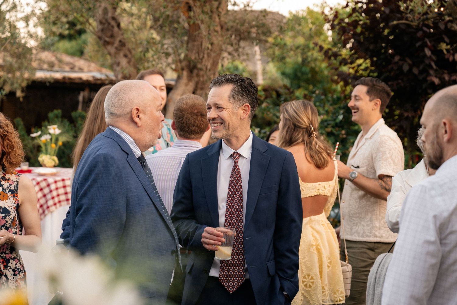 Two elegantly dressed men smiling and conversing at an outdoor garden wedding reception.