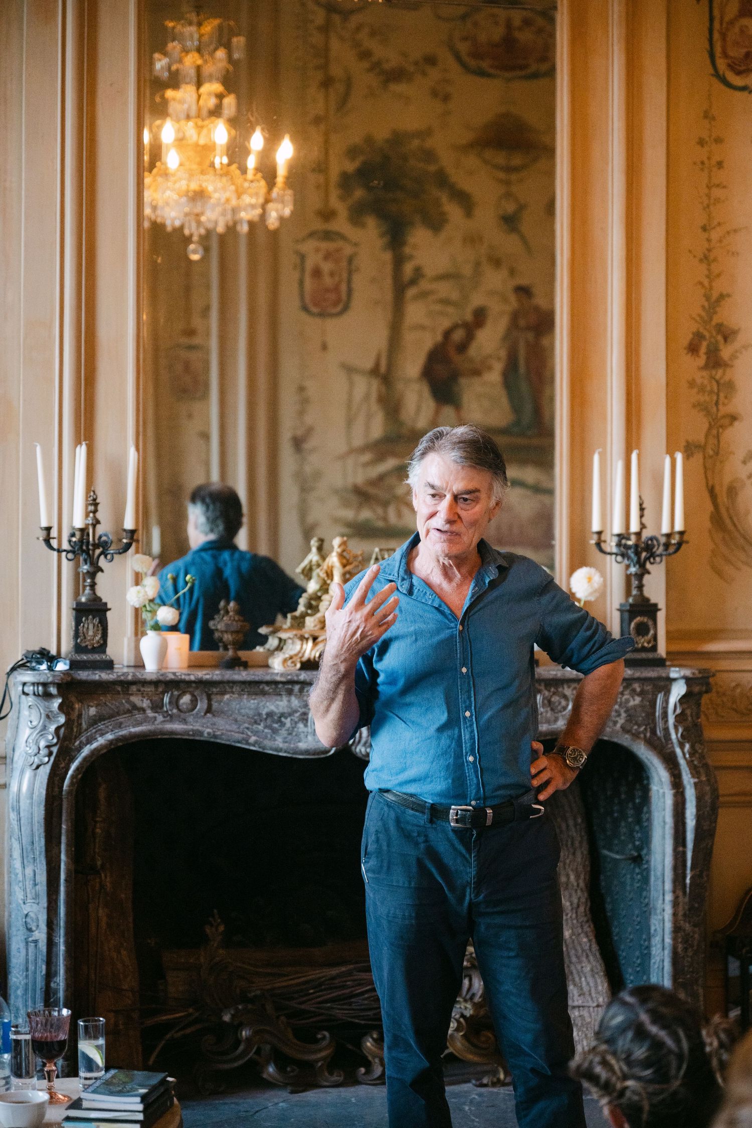 Man gestures while speaking near an ornate marble fireplace with candelabras in a historic chateau.