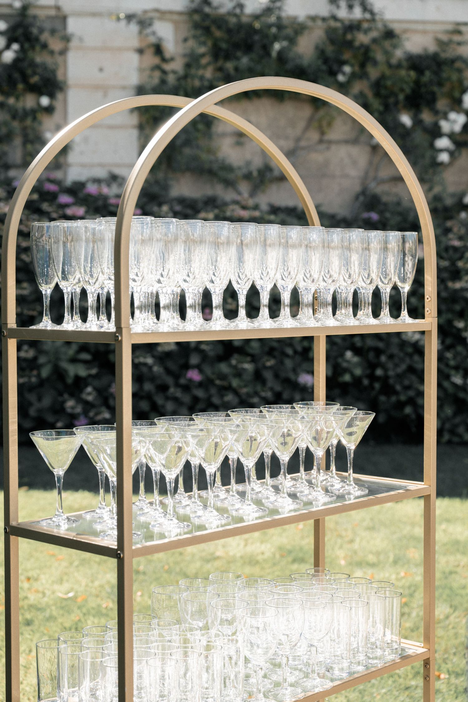 Gold arched display shelf with multiple tiers of crystal champagne flutes and martini glasses at an elegant outdoor event.