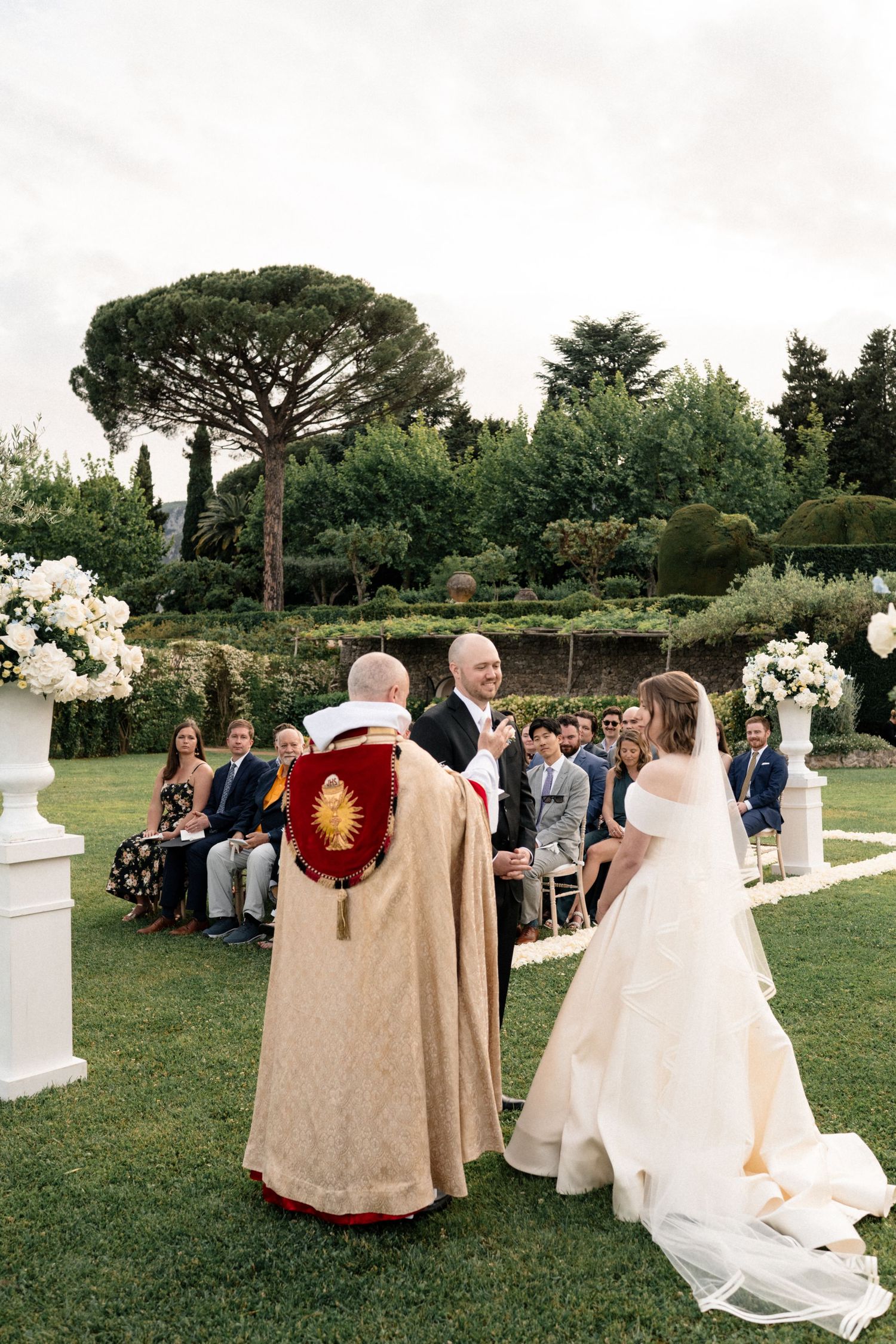 Bride and groom stand before officiant in golden vestments during outdoor garden wedding ceremony in Italy.