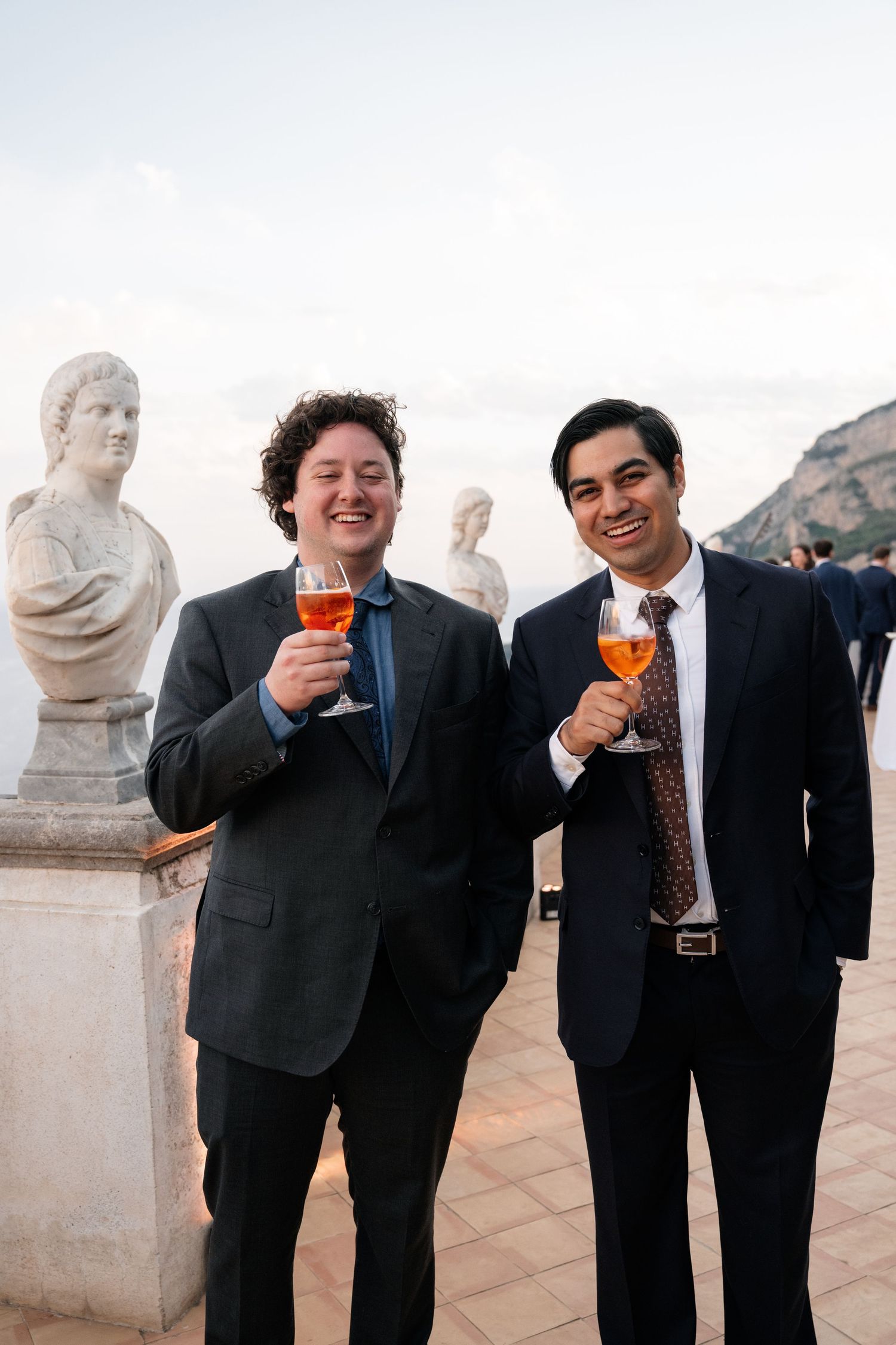 Two male wedding guests toast with aperol spritz beside classical stone busts outdoors.