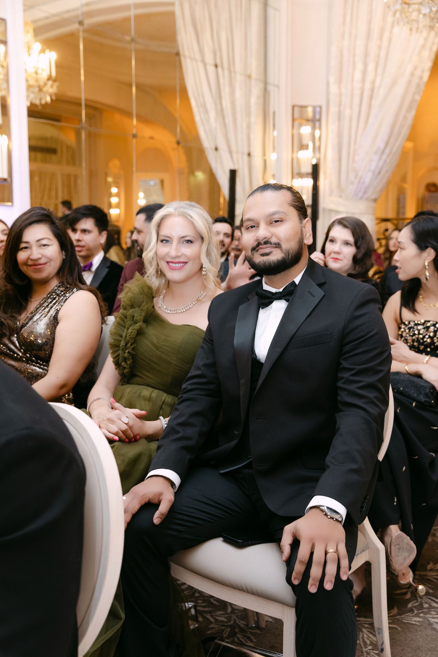Couple in formal attire, woman in olive green dress and man in black tuxedo, smiling at a wedding ceremony.