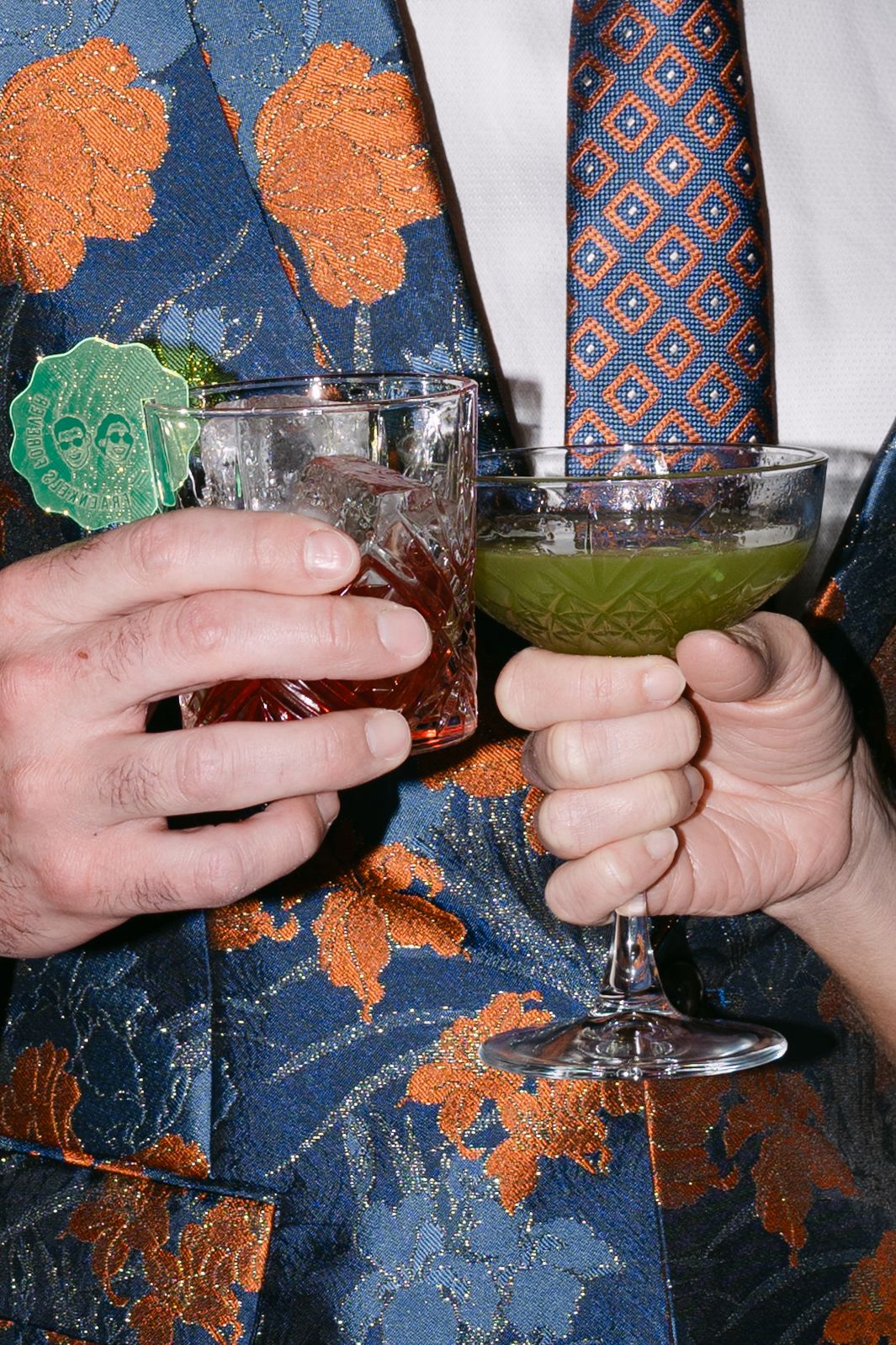 Man in floral blue and orange suit holding two cocktails, red drink and green drink, at event.