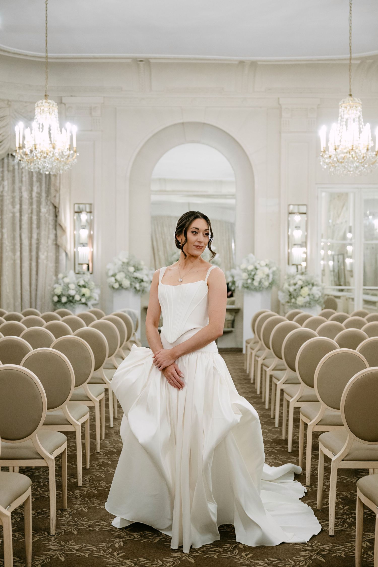 Bride in white satin gown with train posing between rows of gold chairs in crystal chandelier ballroom.
