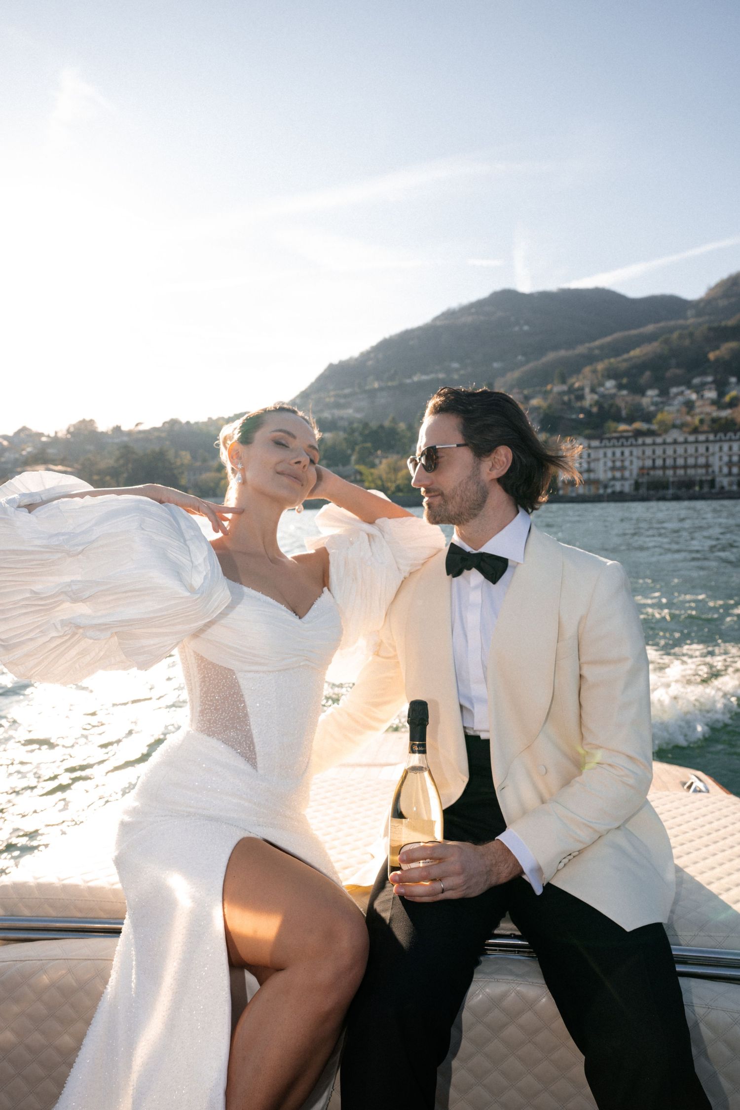Bride in white gown with flowing sleeves sits with groom holding champagne on luxury Lake Como boat.