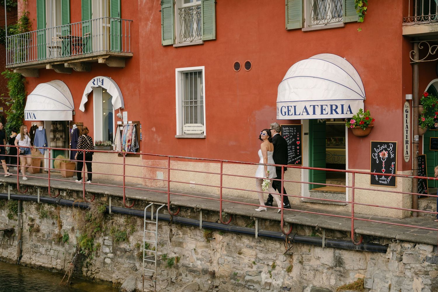 Tourists stroll along a waterfront promenade past a red gelateria building in a charming Italian lakeside town.