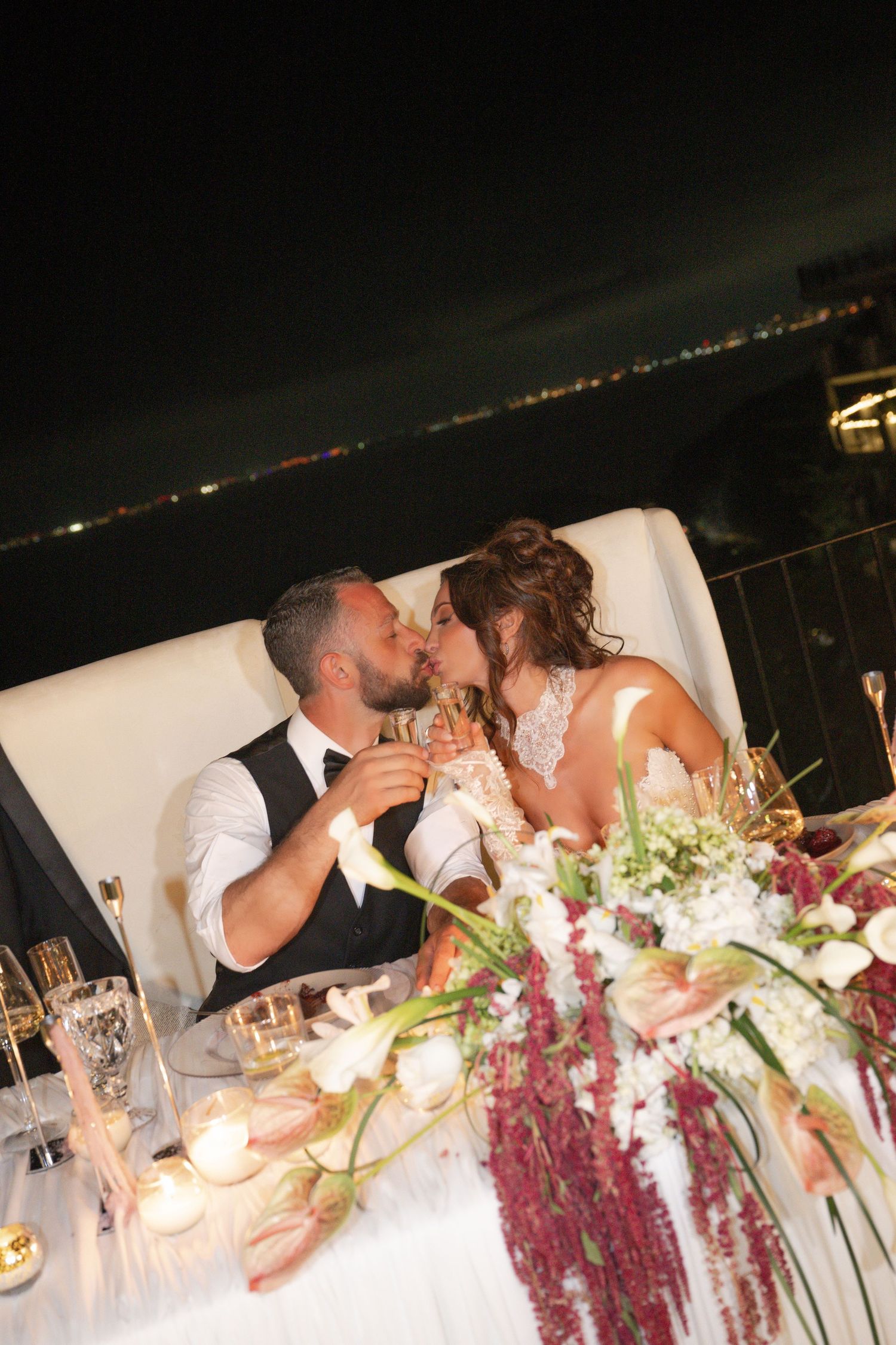 Bride and groom share a romantic kiss at their sweetheart table adorned with floral arrangements and candles at night.