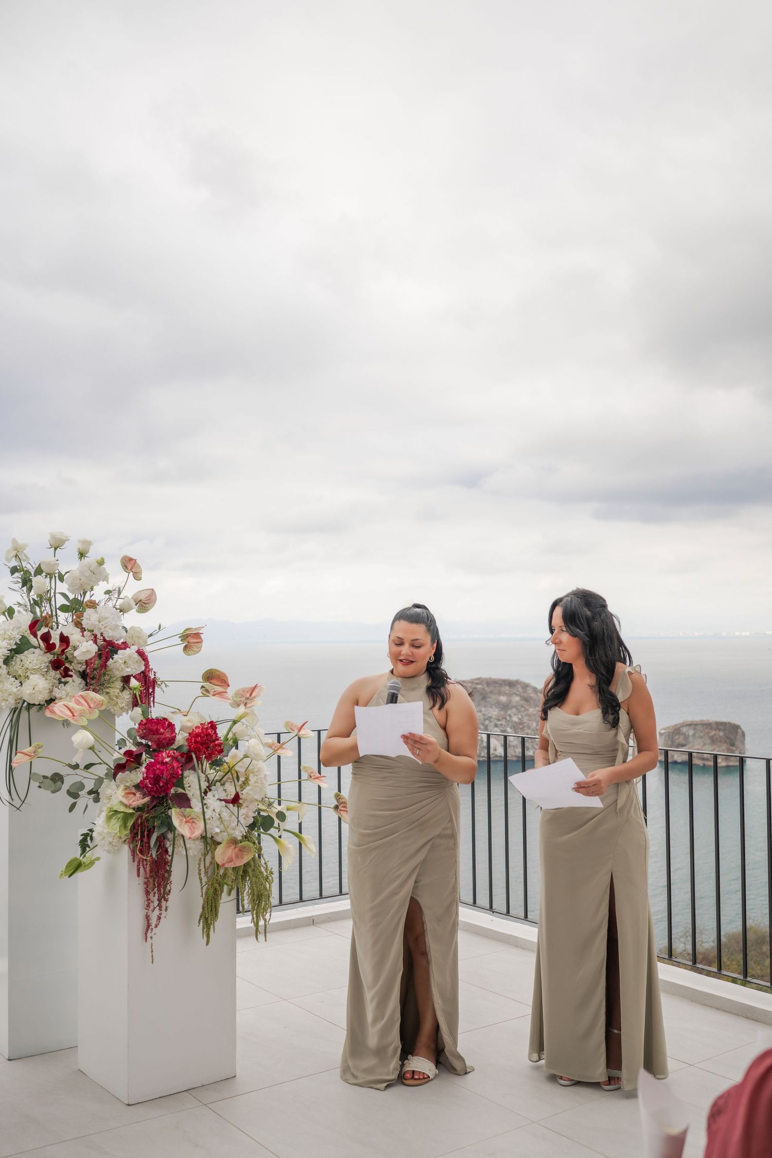Two bridesmaids in taupe gowns deliver readings at outdoor wedding ceremony with ocean view backdrop.