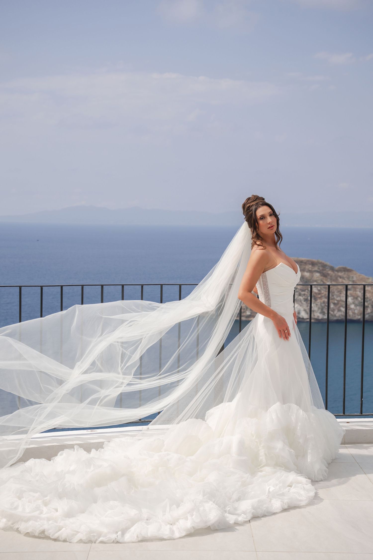 Bride in flowing white gown with dramatic cathedral veil billowing on coastal terrace.