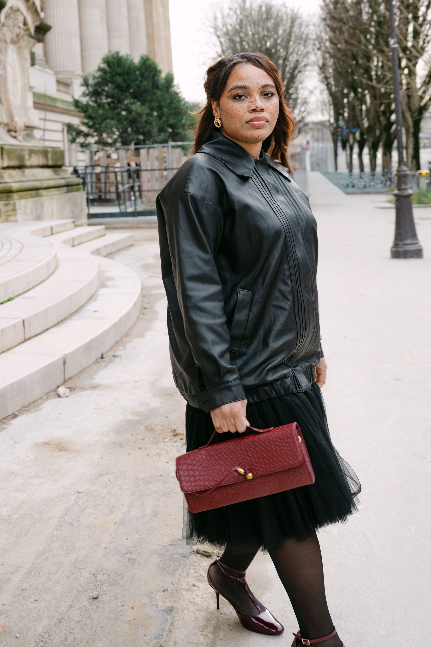 Stylish woman holding burgundy handbag wearing black leather jacket and tulle skirt outside classic Paris building.