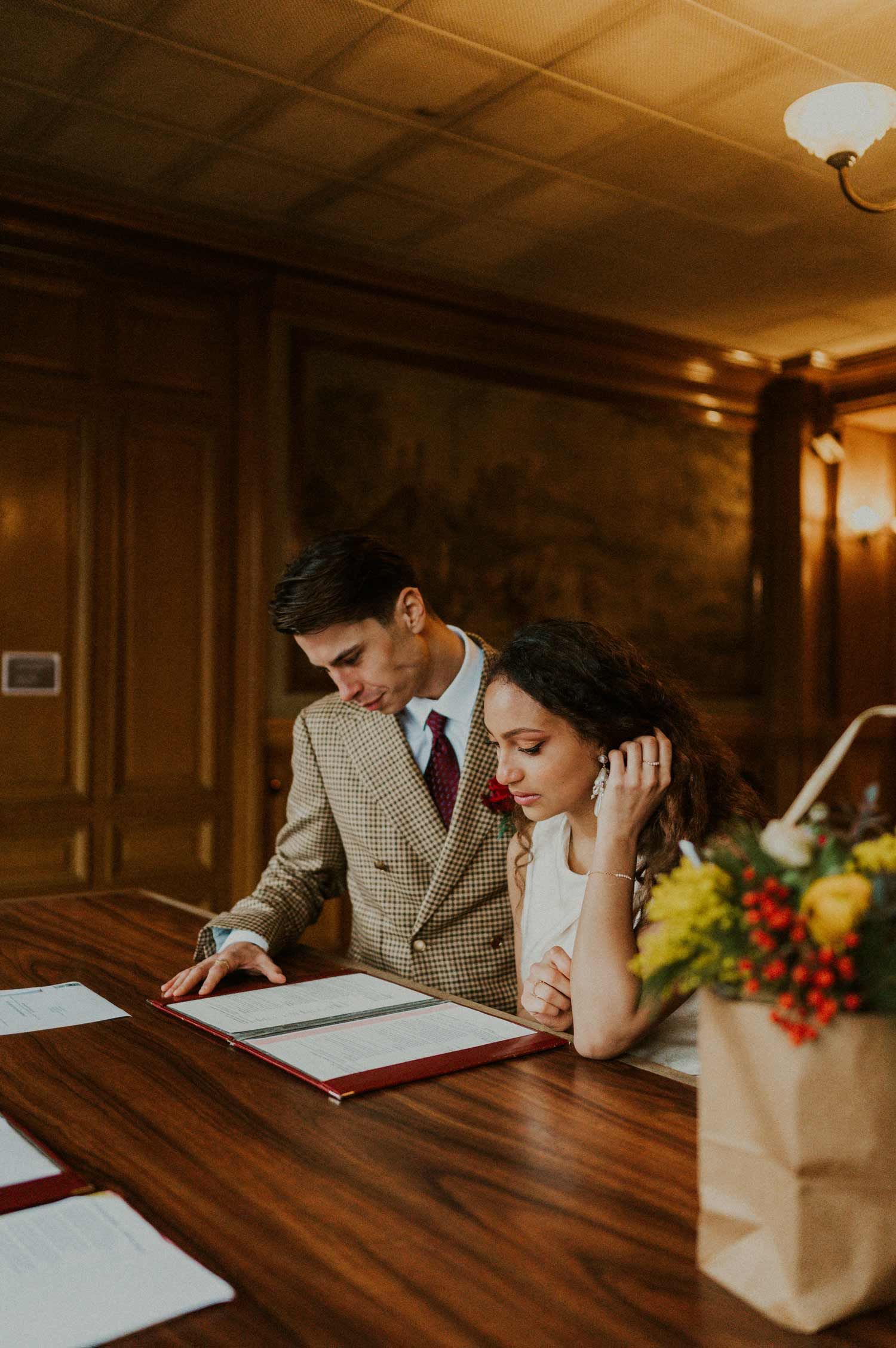 A couple in formal attire signing documents at a wooden table with a floral arrangement nearby.