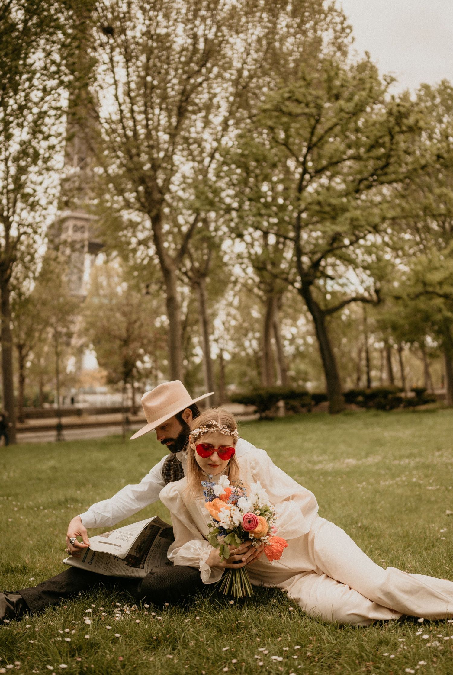 Romantic couple lounging on grass in vintage attire with bouquet and Eiffel Tower visible in background.
