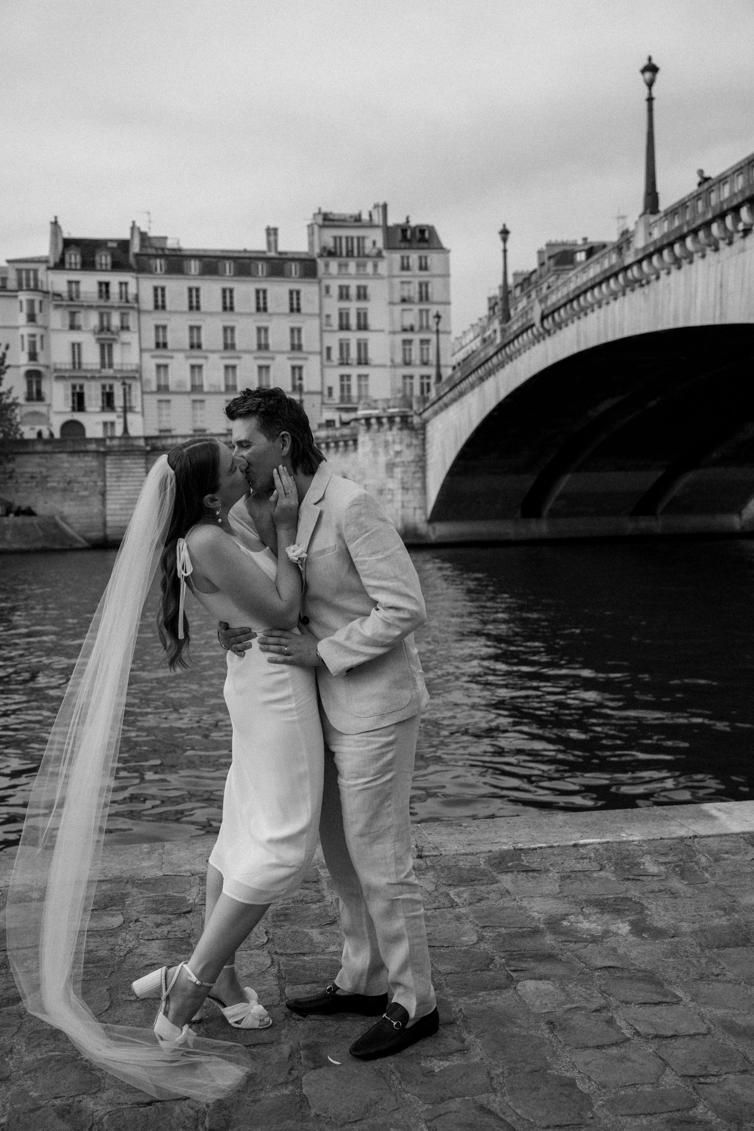 A romantic couple in wedding attire share intimate moments along the Seine River in Paris with historic bridges in view.
