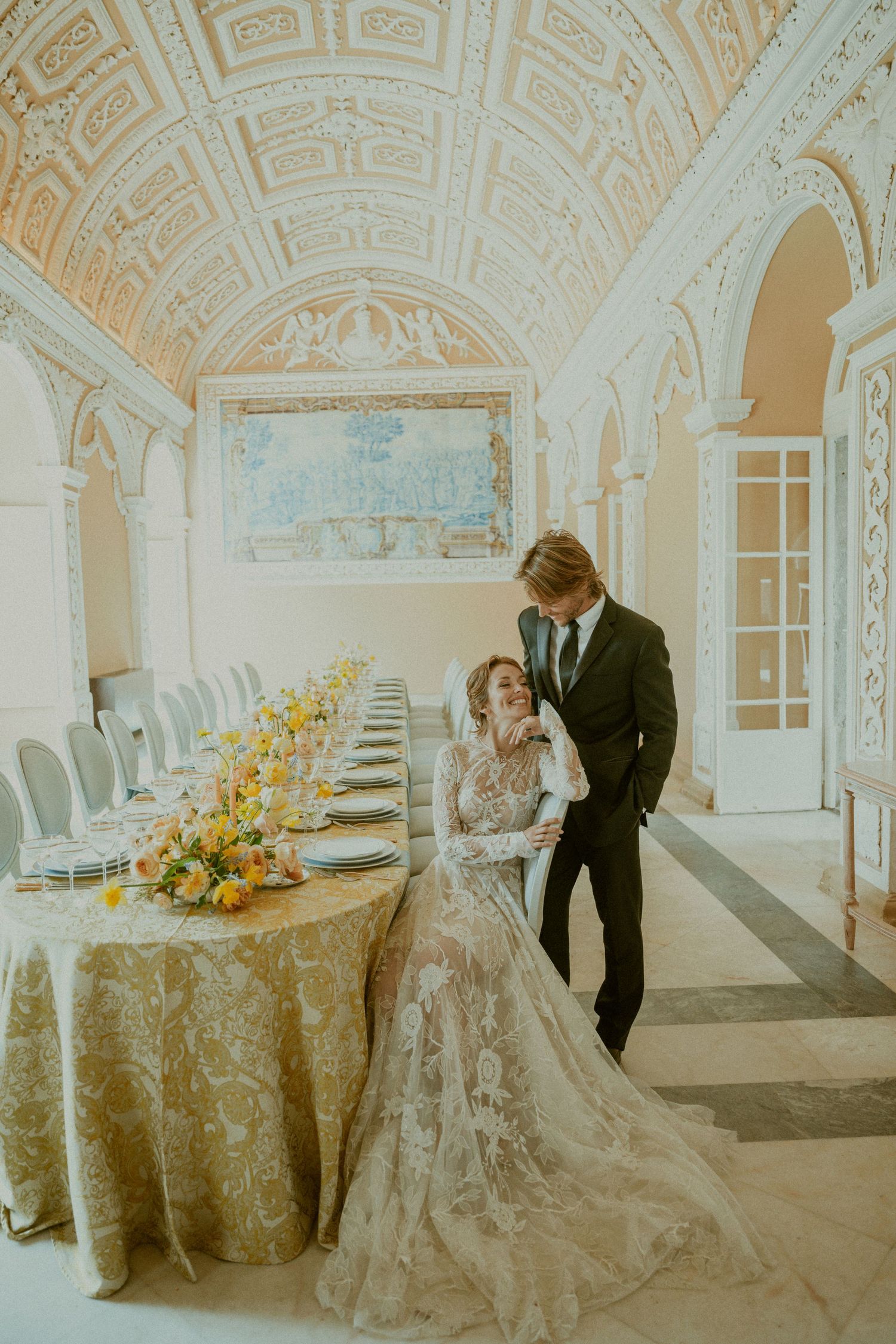 Elegant wedding couple posing in ornate ballroom with long table decorated with yellow floral centerpieces.