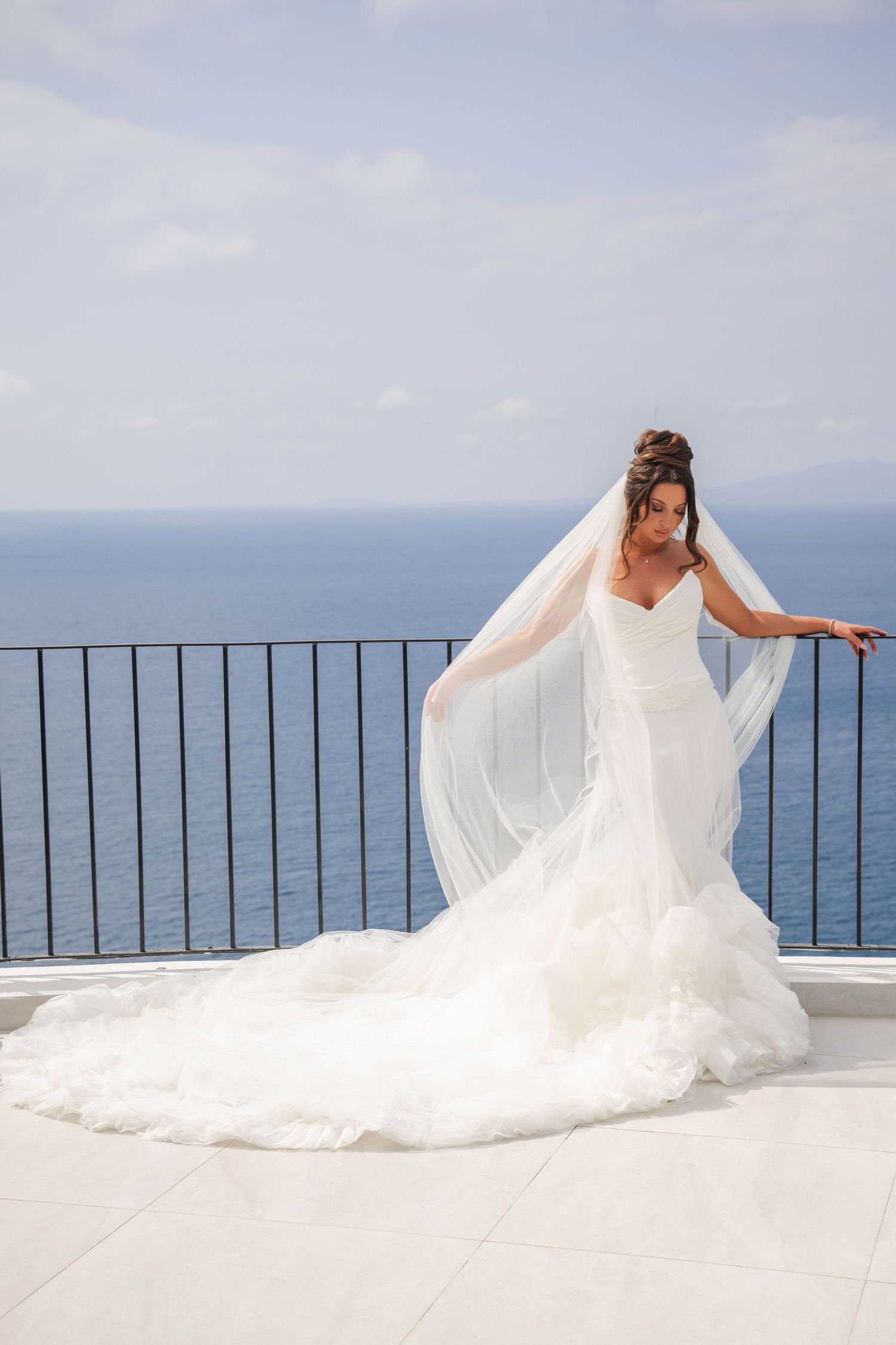 Elegant bride in flowing white wedding dress and veil stands on coastal balcony overlooking the sea.