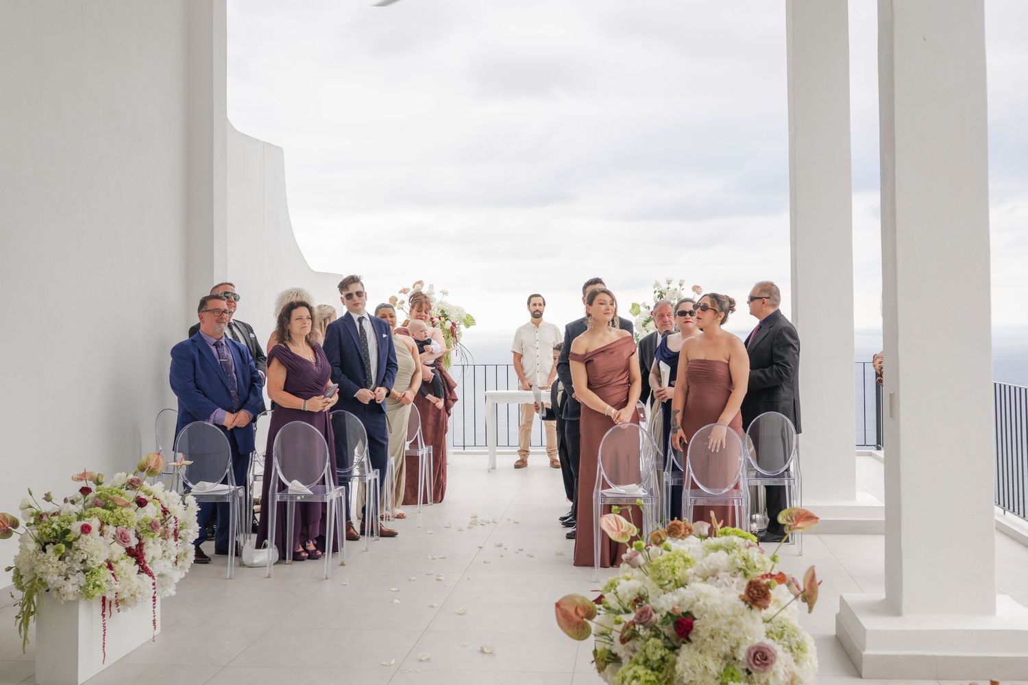 Outdoor wedding ceremony on a white terrace with guests standing along the aisle, floral arrangements, and ocean views.