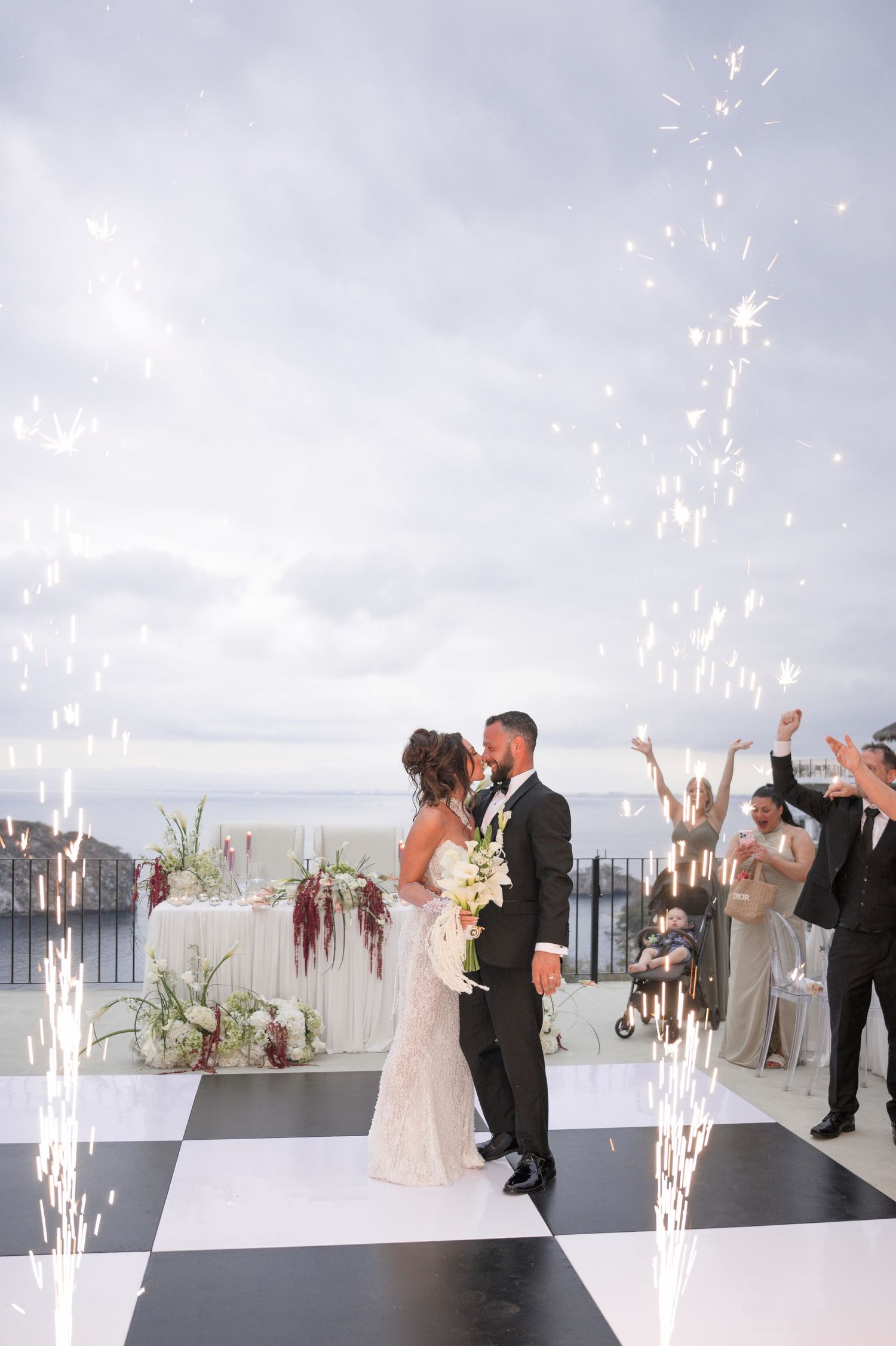 Bride and groom share first kiss on checkered dance floor with sparklers and ocean backdrop at elegant outdoor wedding.