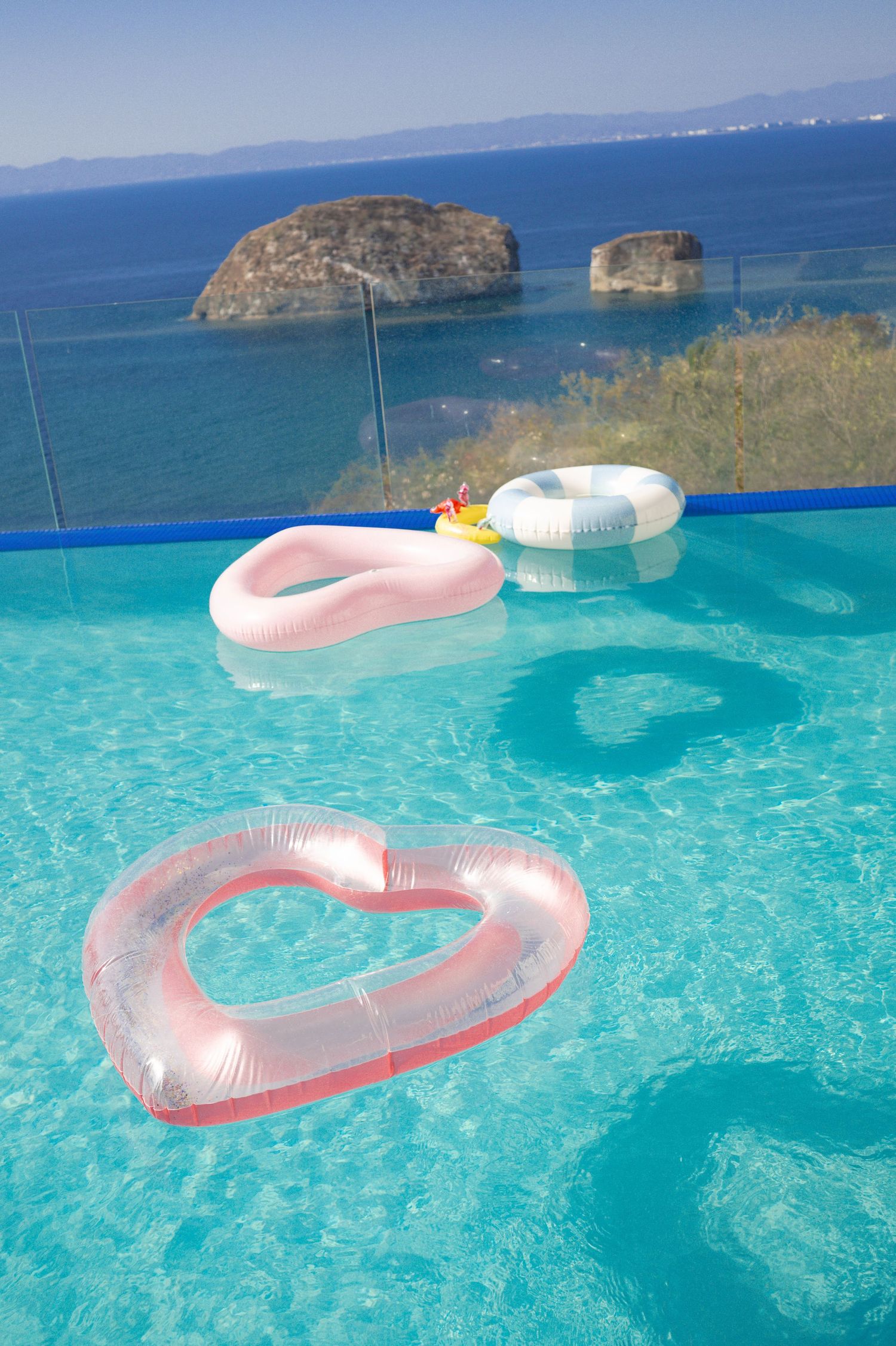 Colorful inflatable pool floats drift in a turquoise infinity pool overlooking a scenic ocean view.