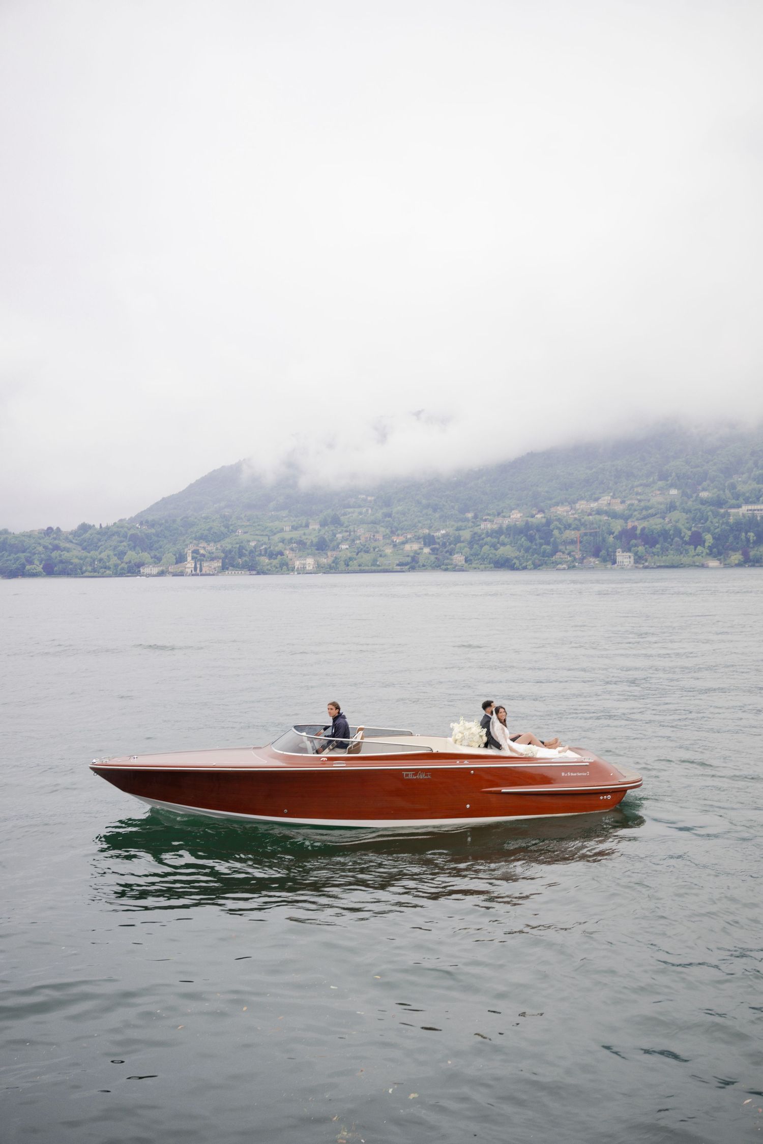 Bride and groom riding a classic wooden speedboat across a misty Italian lake.