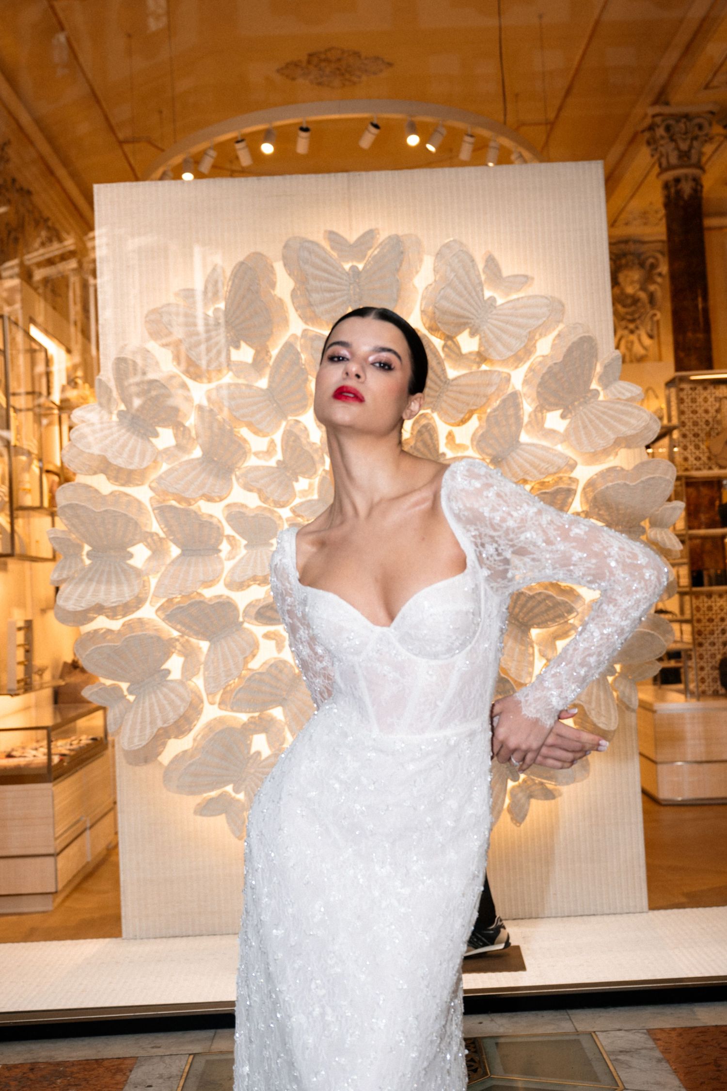 A woman in a fitted white lace long-sleeve wedding gown poses before a glowing butterfly wall display.
