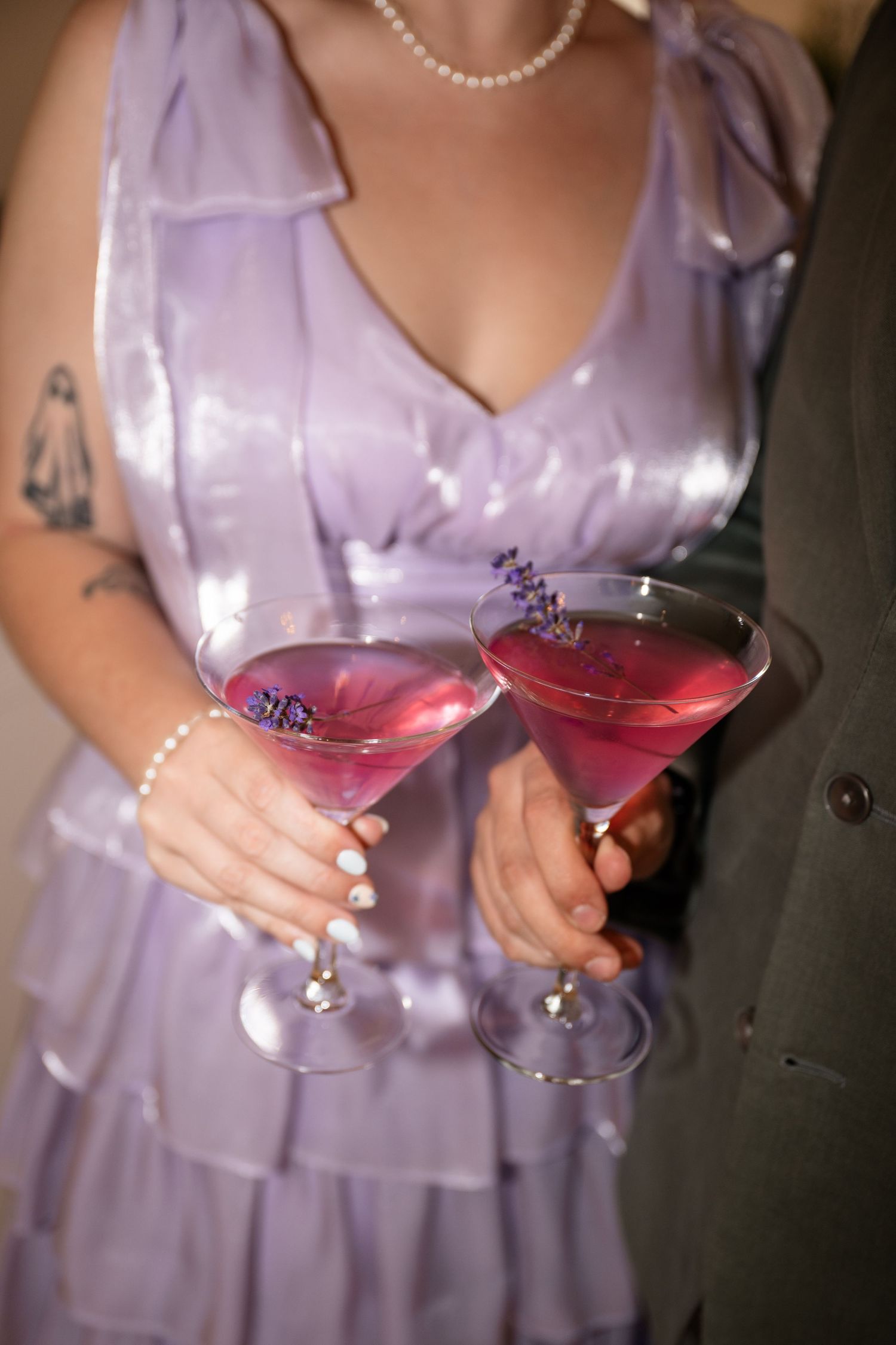 Two pink cocktails in martini glasses held by someone wearing a lavender dress with pearl accessories.