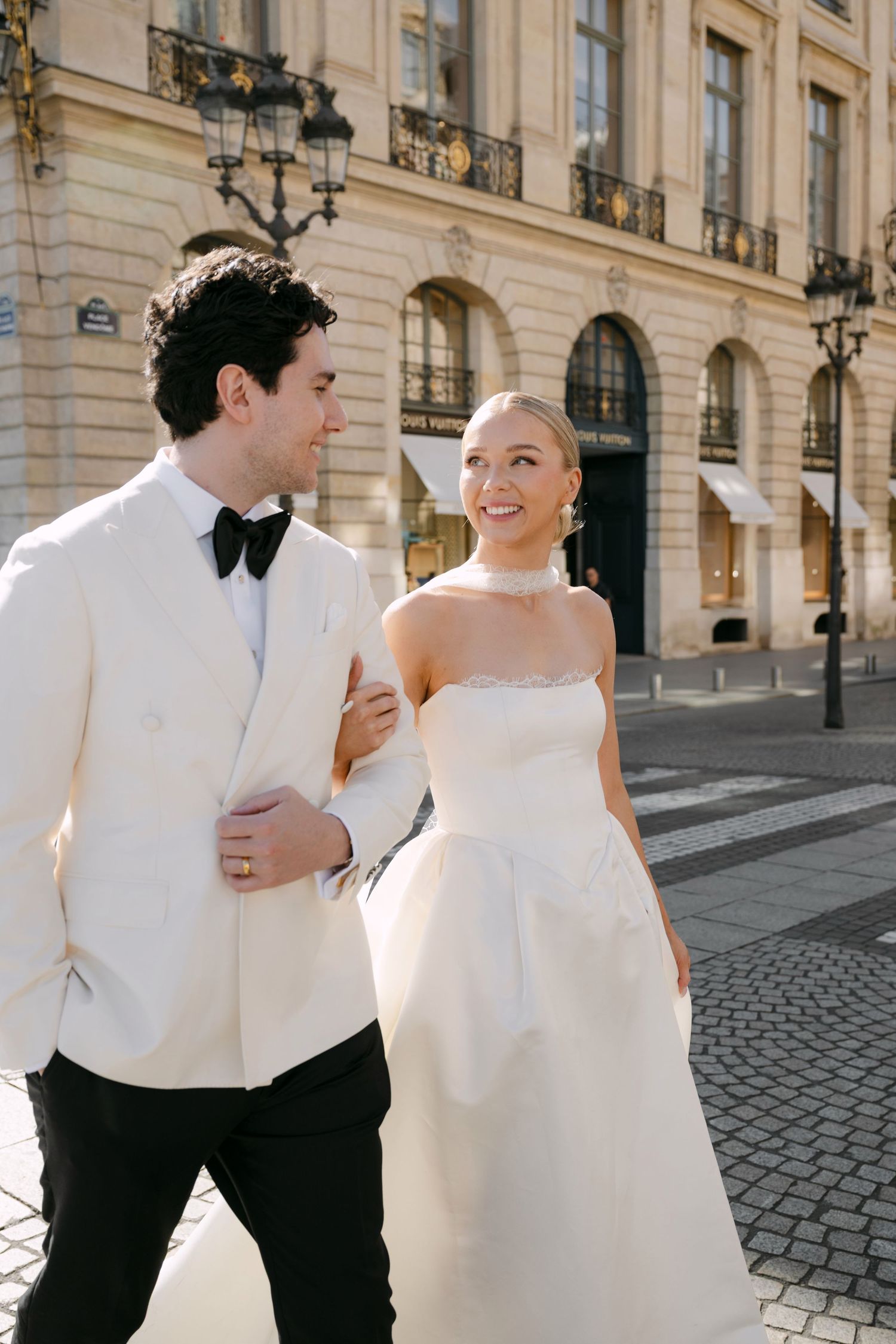 A couple in wedding attire walks and laughs together on a cobblestone street in front of elegant European buildings.
