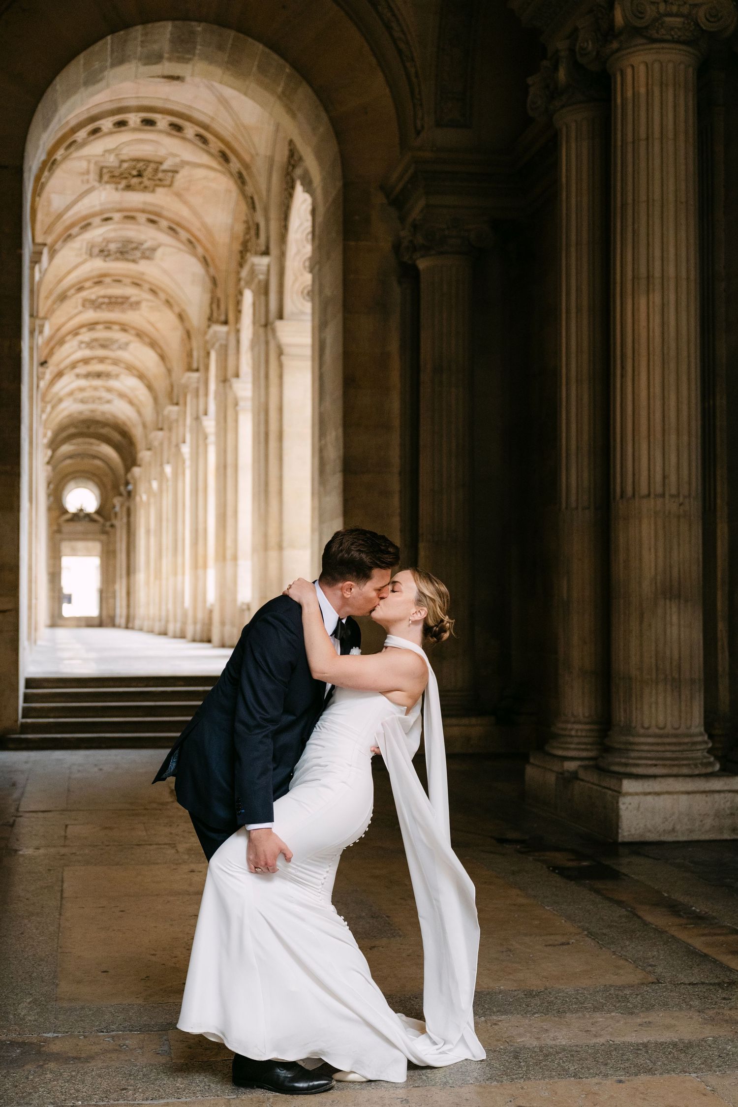 Romantic wedding photo in an elegant arched hallway with classical columns and stone architecture.