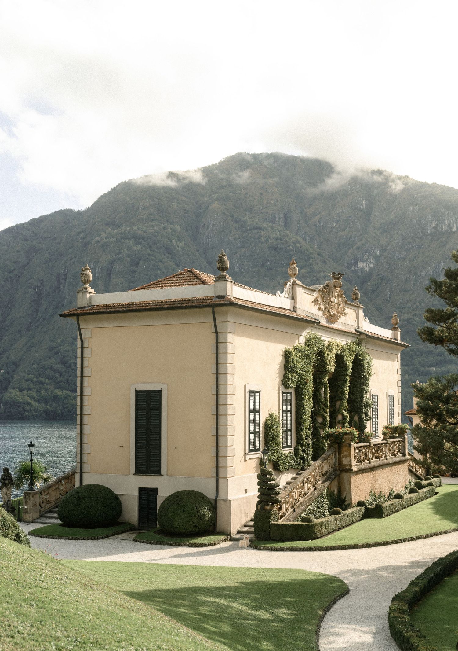 A white neoclassical villa with decorative statues sits beside a lake with mountains in the background.